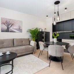 Kreator Designu - Jasne wnętrze salonu z aneksem kuchennym: szara sofa z poduszką, czarny stolik kawowy z dwoma filiżankami, okrągły stół z czterema krzesłami, zabudowa kuchenna z czarnymi frontami i jasnymi...