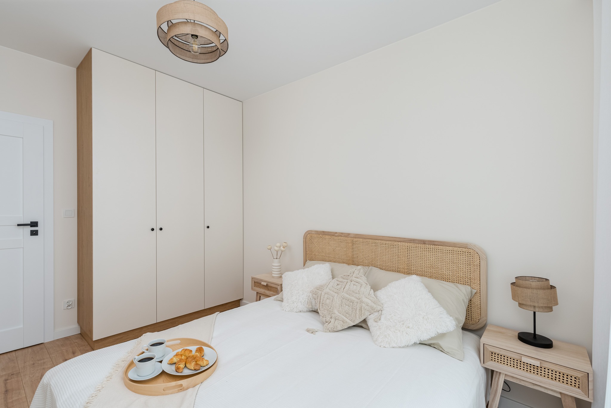 Jasna sypialnia w stylu boho z rattanowym wezgłowiem łóżka, beżowymi poduszkami, drewnianą szafką nocną z lampką i śniadaniem na tacy