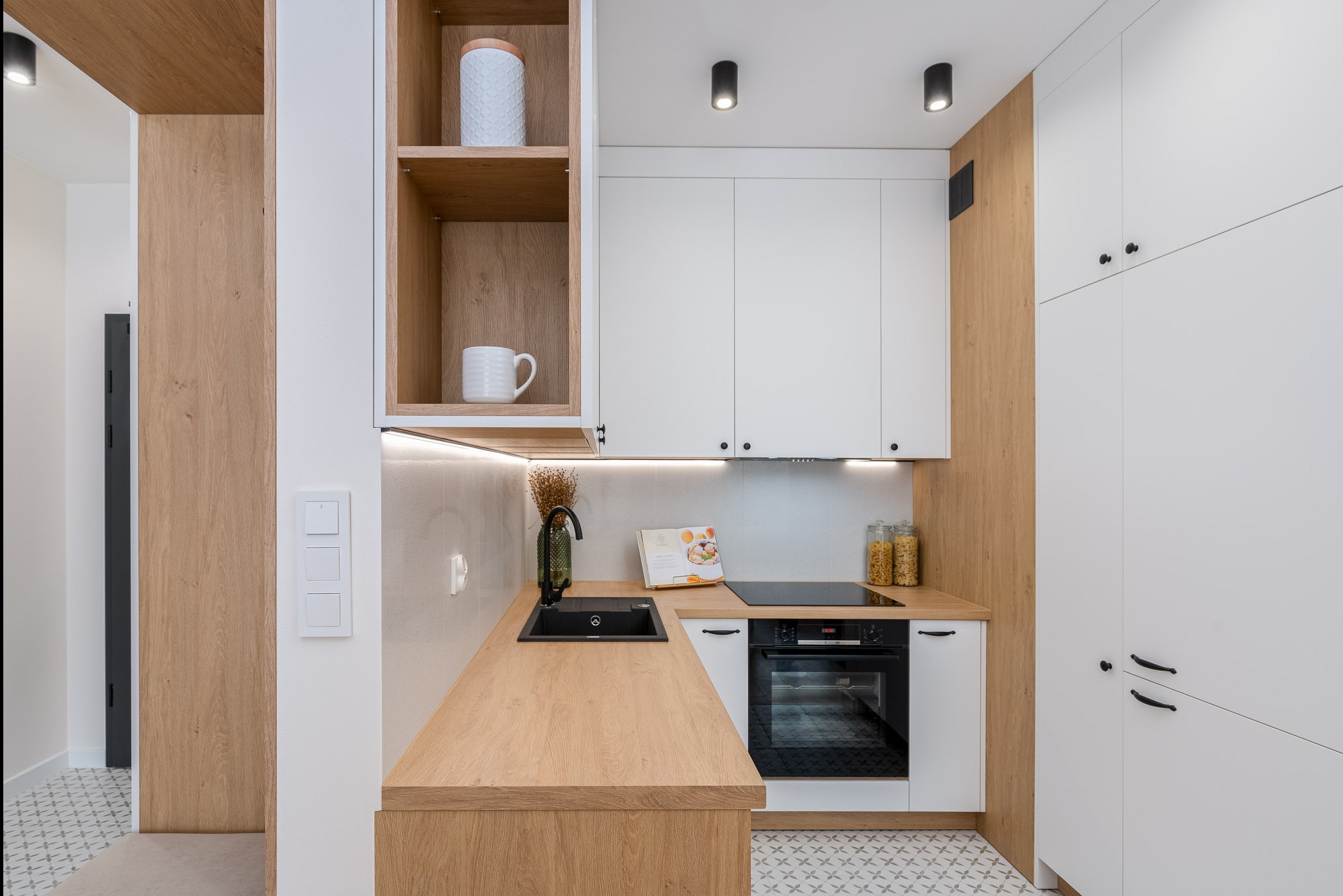 Nowoczesna kuchnia z białymi szafkami, drewnianym blatem i czarną armaturą, oświetlona punktowymi lampami sufitowymi, z widoczną książką kucharską i dekoracyjnymi słoikami.