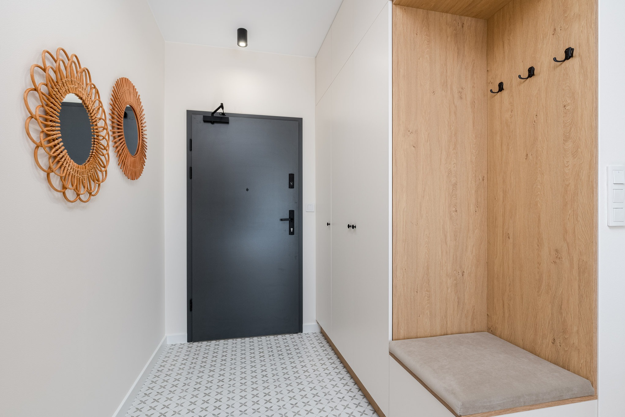 Nowoczesny przedpokój z szarą, antywłamaniową  drzwi wejściowymi,  dekoracyjnymi lustrami w rattanowych ramach, szafą w zabudowie z siedziskiem wykończonym drewnem oraz wzorzystą podłogą.