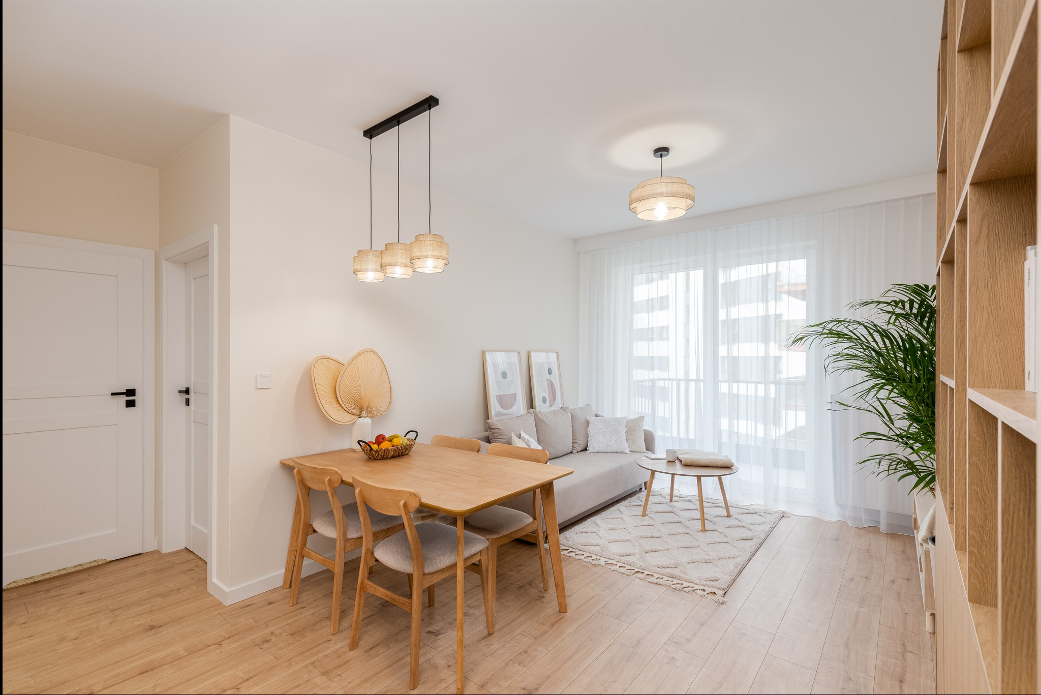 Jasne, nowocześnie urządzone wnętrze mieszkania z drewnianym stołem, krzesłami, sofą i regałem, oświetlone wiszącymi lampami z abażurami z naturalnego materiału.