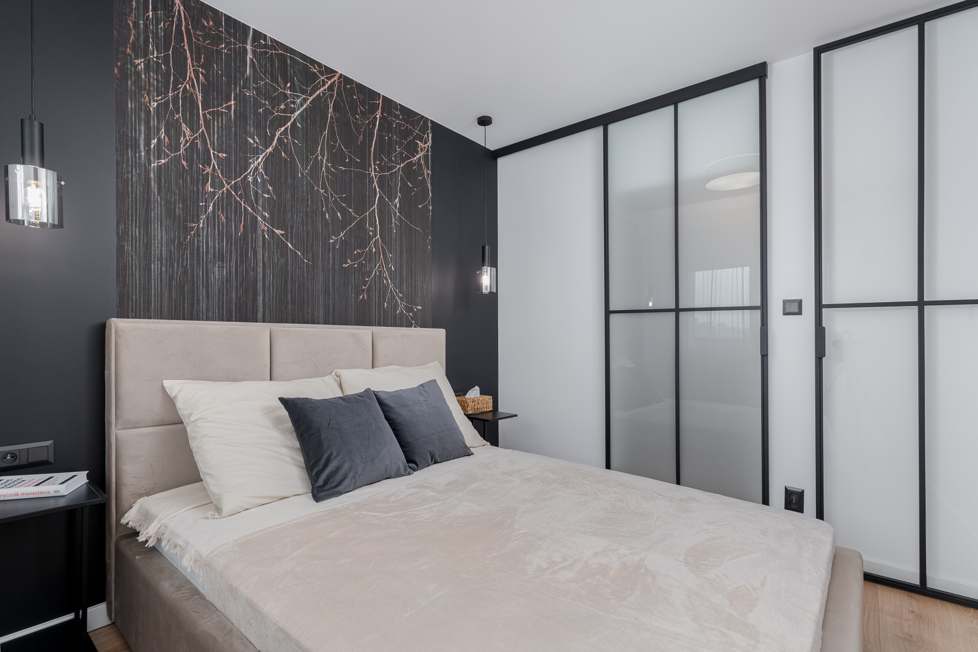 Nowoczesna sypialnia z beżowym łóżkiem, szarą ścianą z motywem drzewa i przesuwnymi drzwiami z czarnymi szprosami.