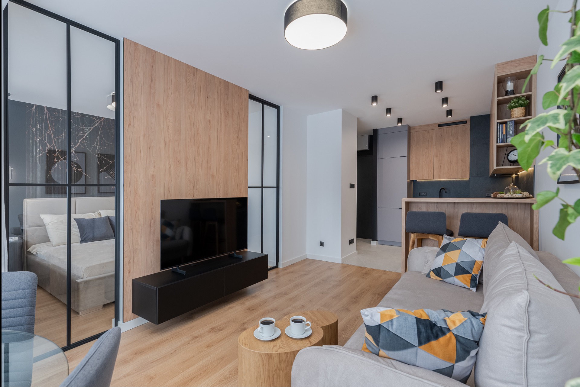Nowoczesne wnętrze: salon z aneksem kuchennym, drewniana ściana z telewizorem, przeszklona ściana oddzielająca sypialnię, minimalistyczne oświetlenie punktowe, kawa na drewnianym stoliku kawowym.