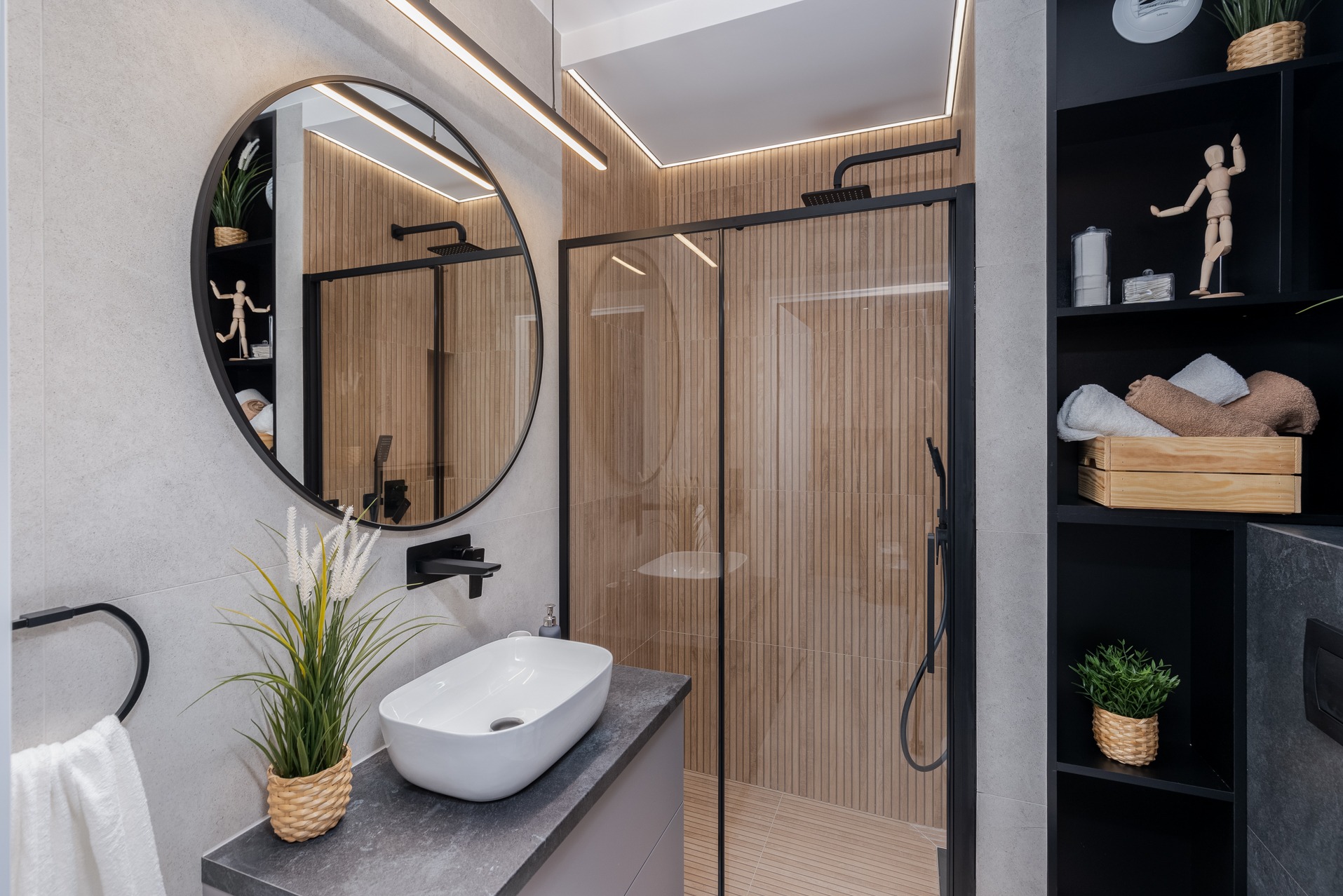 Nowoczesna łazienka z kabiną prysznicową z czarnymi ramami, okrągłym lustrem z czarną obwódką odbijającym wnętrze kabiny, umywalką nablatową i drewnopodobnymi płytkami na ścianach kabiny.
