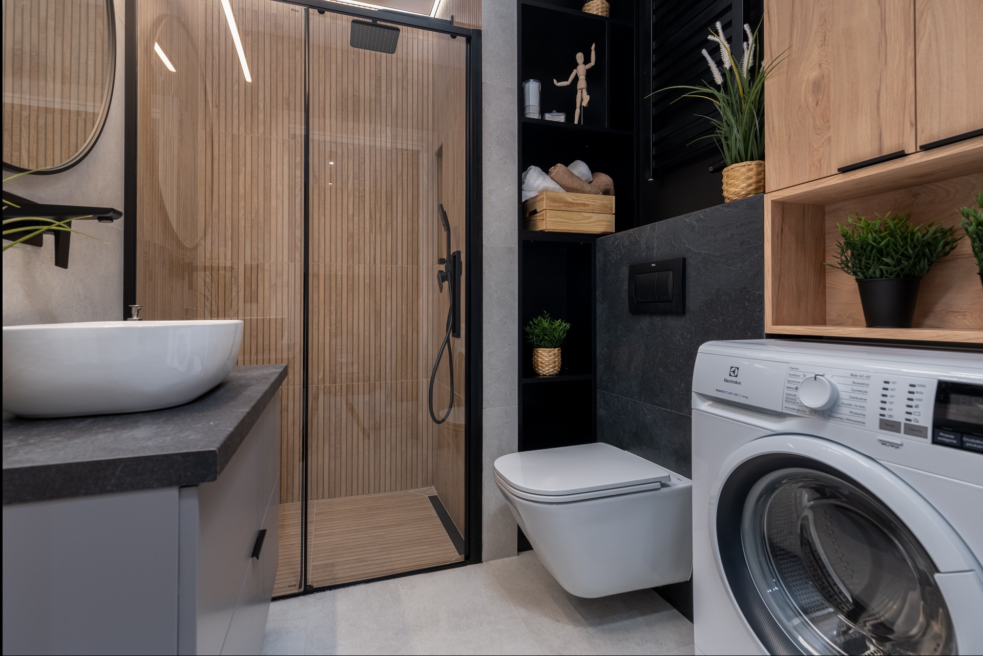 Nowoczesna łazienka z kabiną prysznicową wyłożoną drewnopodobnymi płytkami, umywalką nablatową, wiszącą toaletą, pralką i czarnymi półkami z dekoracjami.
