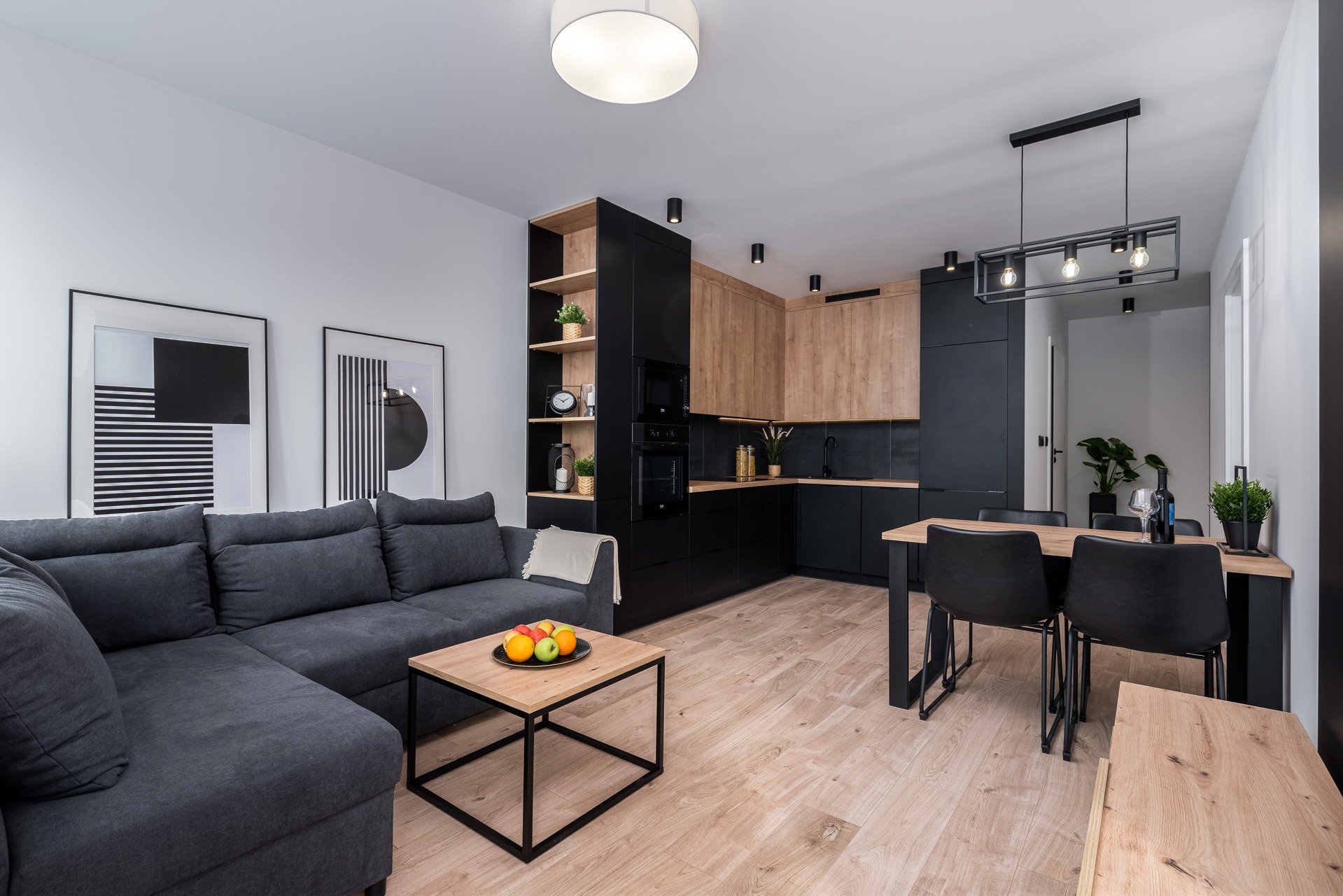 Nowoczesne wnętrze: szary narożnik, drewniany stolik z owocami, czarno-drewniana zabudowa kuchenna, stół z krzesłami, minimalistyczne obrazy na ścianie.