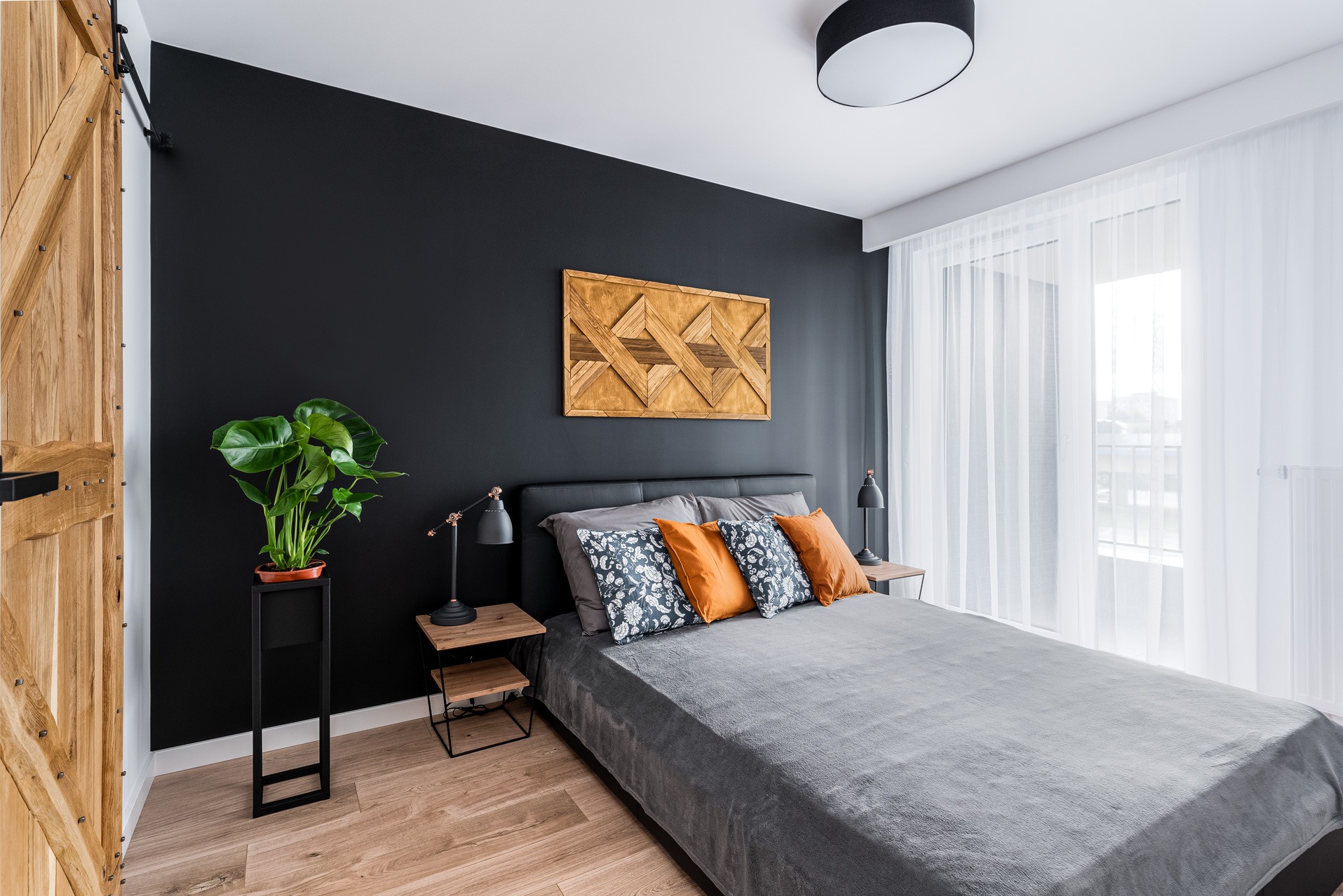 Nowoczesna sypialnia z czarną ścianą, drewnianym panelem dekoracyjnym, łóżkiem z szarą narzutą, pomarańczowymi i wzorzystymi poduszkami, lampkami nocnymi oraz drewnianymi drzwiami przesuwnymi typu...