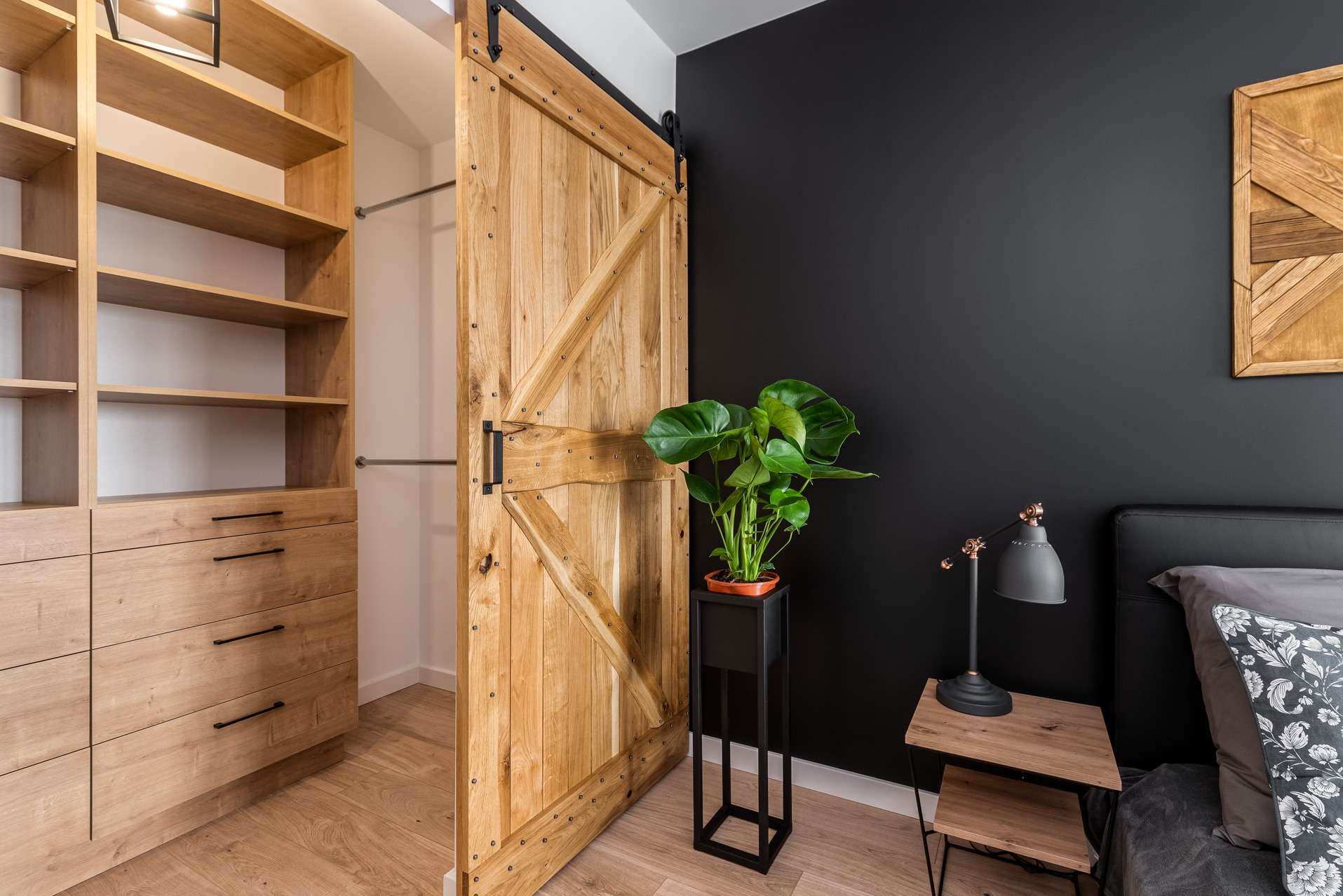 Drewniana, przesuwna brama do garderoby z widoczną zawartością szaf, w tle czarna ściana z drewnianą dekoracją, obok stolik nocny z lampką i rośliną w doniczce.