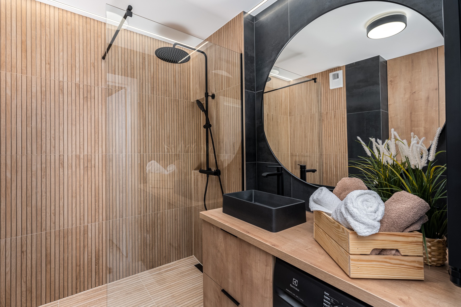 Nowoczesna łazienka z drewnopodobnymi płytkami ściennymi, czarną armaturą, okrągłym lustrem i drewnianym blatem z czarną umywalką oraz dekoracyjnym pudełkiem z ręcznikami.