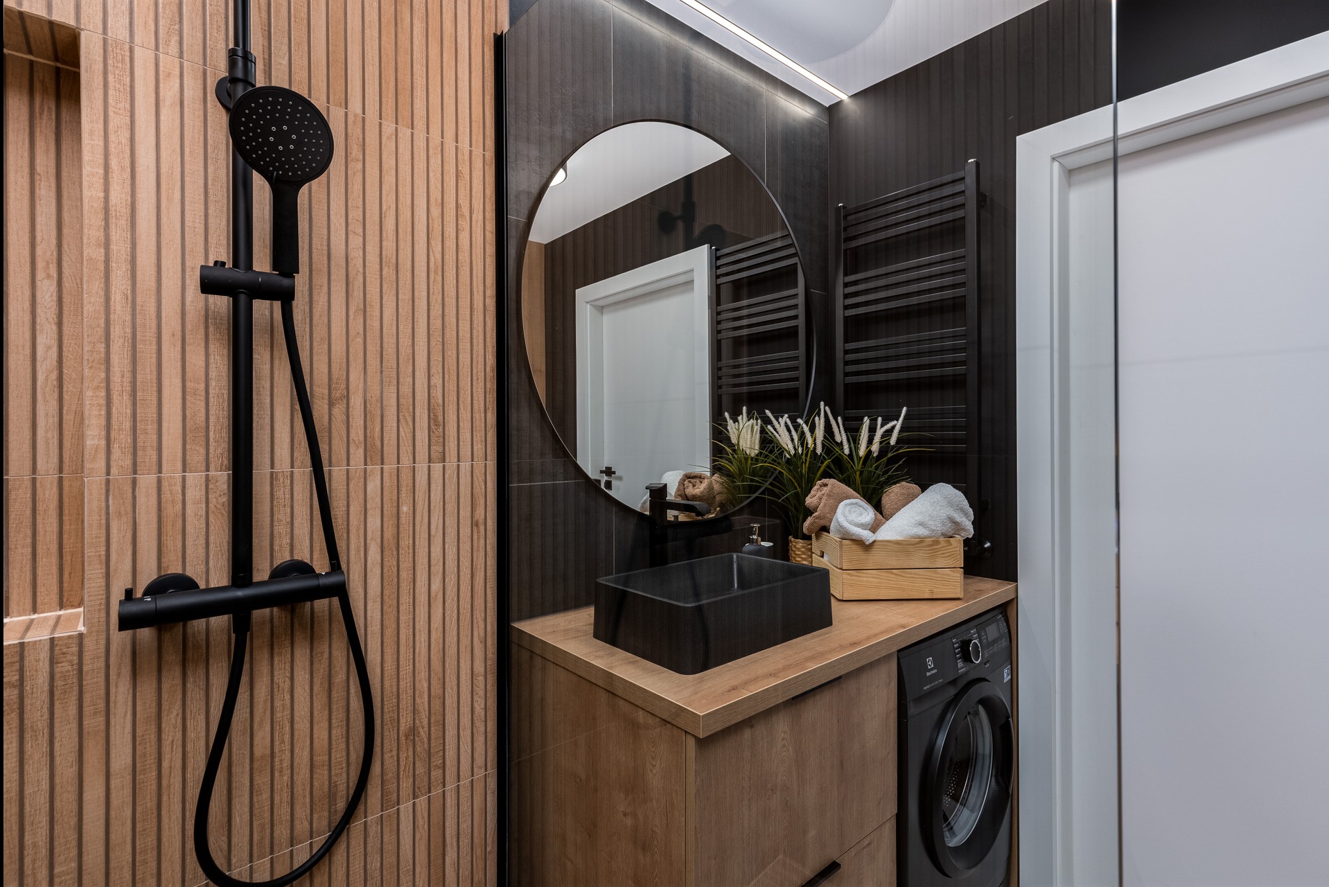 Nowoczesna łazienka z drewnopodobnymi płytkami, czarną armaturą, okrągłym lustrem i pralką pod blatem umywalki.