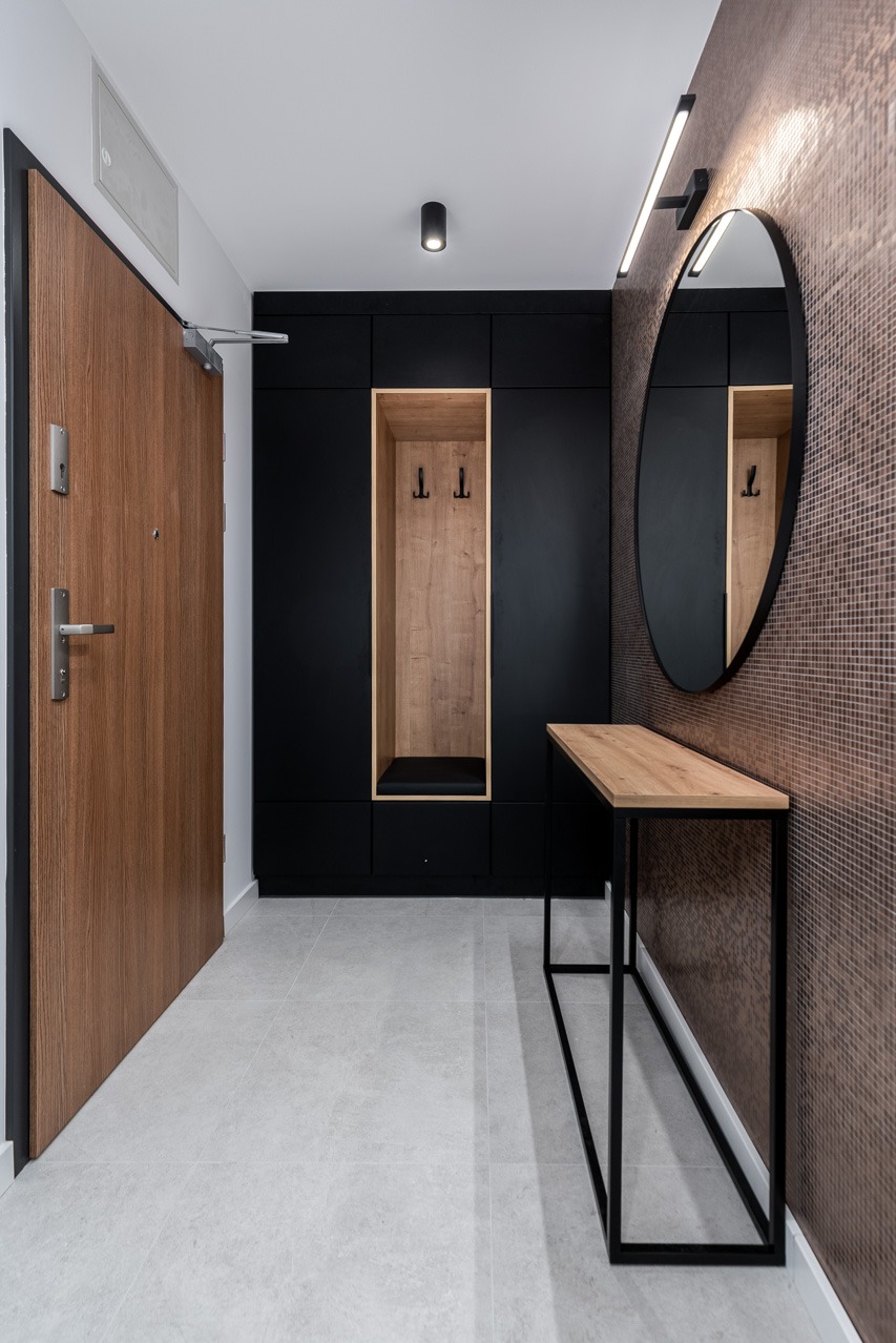 Nowoczesny przedpokój z zabudową w kolorze czarnym i drewnianymi akcentami, okrągłe lustro na mozaikowej ścianie, drewniane drzwi wejściowe.