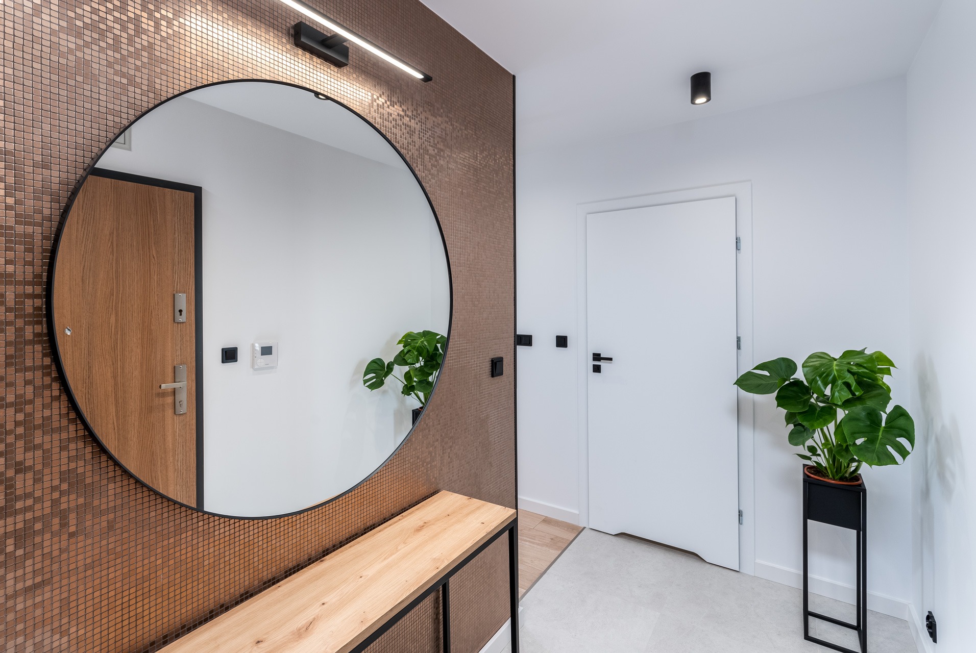 Nowoczesny przedpokój z okrągłym lustrem, mozaikową ścianą w odcieniach brązu, drewnianą ławką i rośliną Monstera w czarnej donicy.