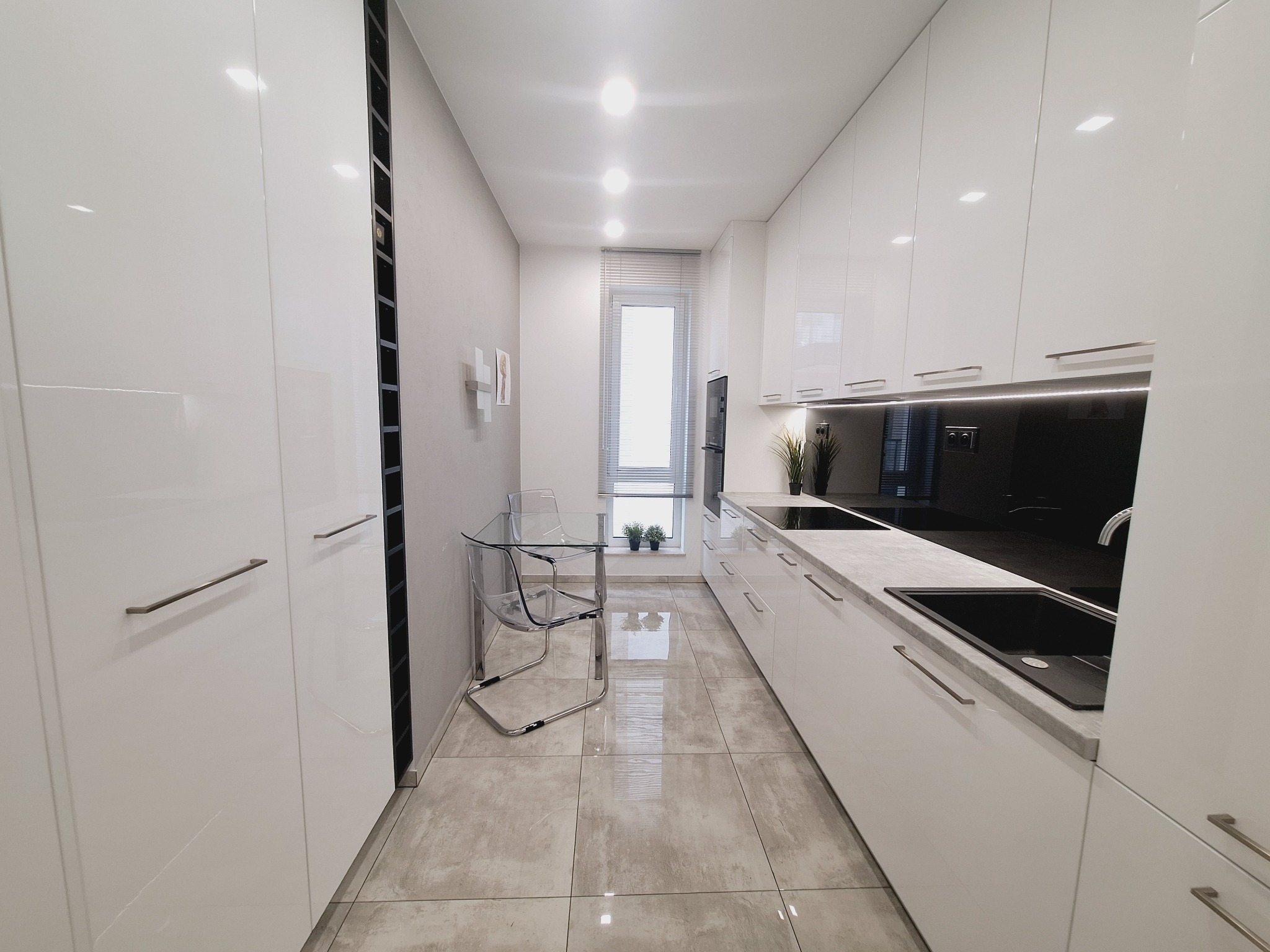 Nowoczesna, minimalistyczna kuchnia w bieli z szarymi akcentami: błyszczące fronty szafek, szary blat, czarny panel nad blatem, przezroczyste krzesła i stół, płytki podłogowe imitujące kamień.