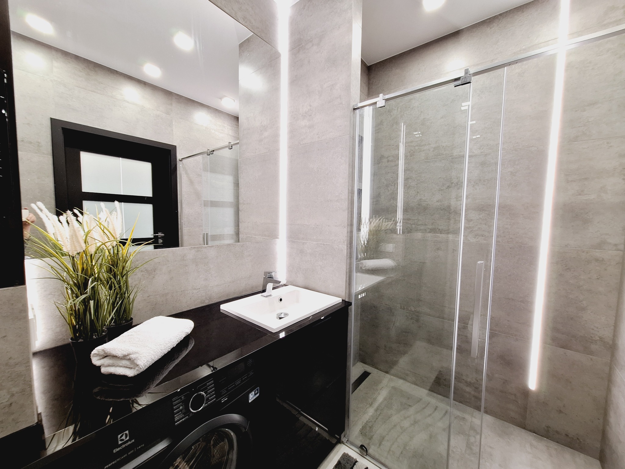 Nowoczesna łazienka w szarościach z kabiną prysznicową ze szklanymi drzwiami przesuwnymi, umywalką nablatową na czarnym blacie, pod którą znajduje się pralka Electrolux oraz dekoracyjną trawą...