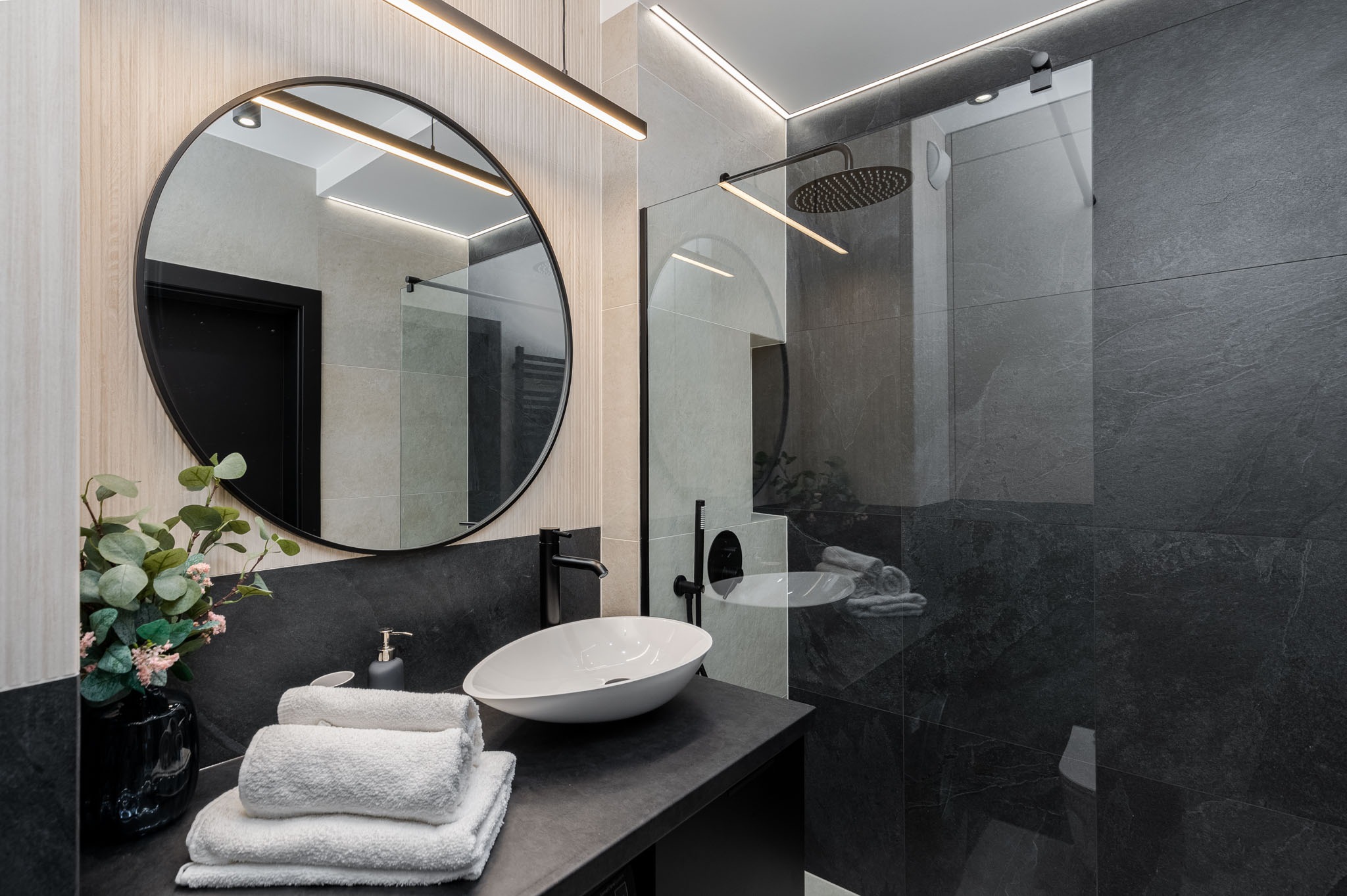 Nowoczesna łazienka z czarnymi płytkami imitującymi kamień, okrągłe lustro z czarną ramą, biała umywalka nablatowa, czarna bateria, szklana kabina prysznicowa typu walk-in i dekoracja roślinna...