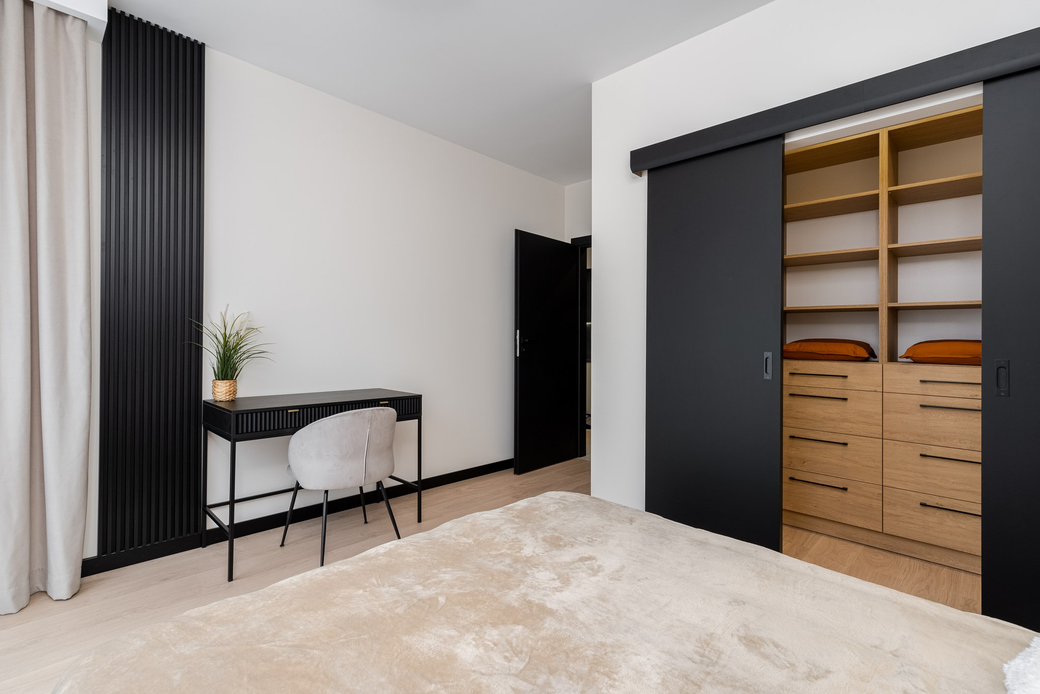 Nowoczesna sypialnia z beżowym łóżkiem, czarnym biurkiem z rośliną, szarą tapicerowaną krzesłem i przesuwną szafą z drewnianymi szufladami i półkami za czarnymi drzwiami.