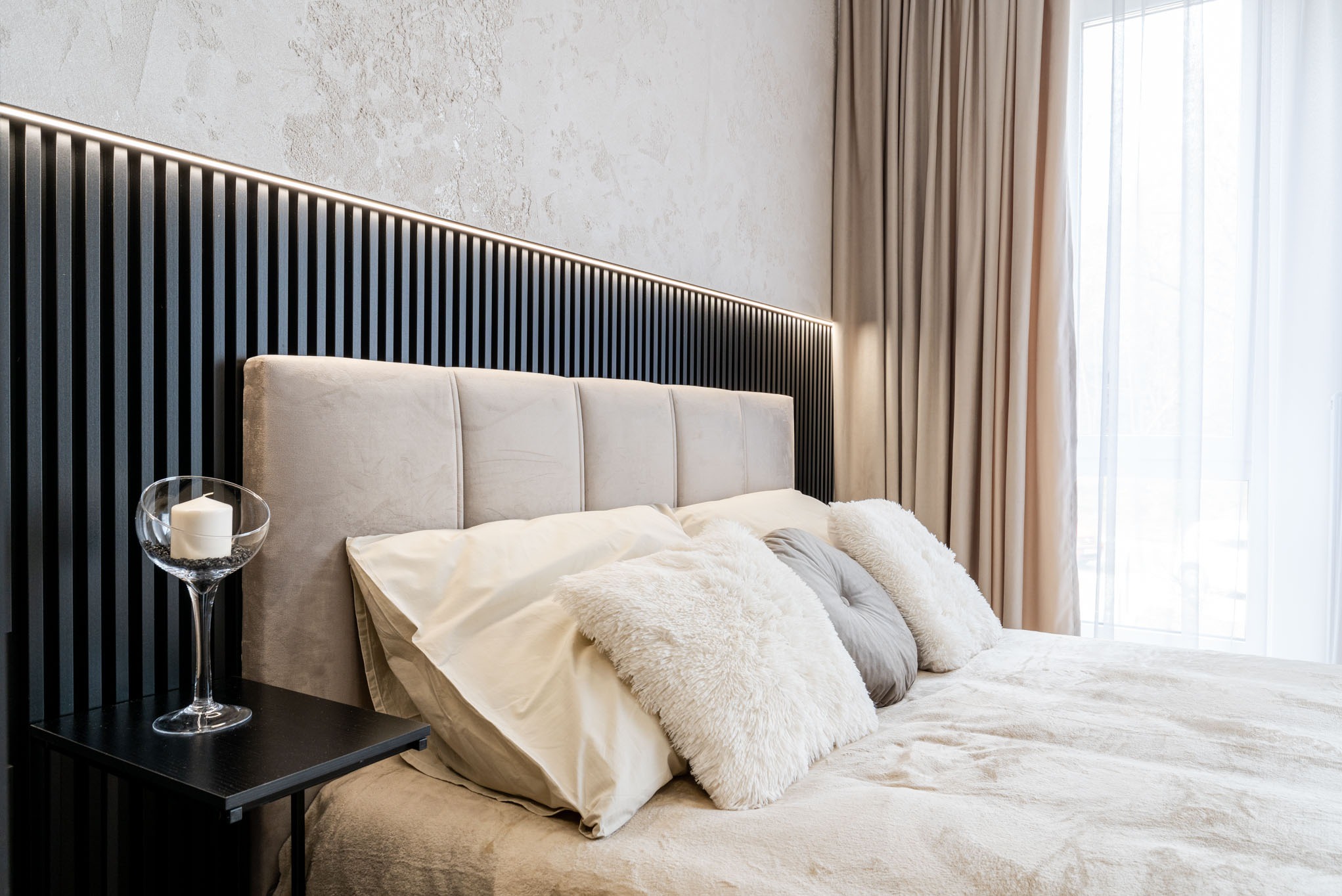 Nowoczesna sypialnia z beżowym tapicerowanym łóżkiem, czarnym zagłówkiem z oświetleniem LED, dekoracyjnymi poduszkami i stolikiem nocnym z kieliszkiem wypełnionym dekoracyjnym żwirkiem i świecą