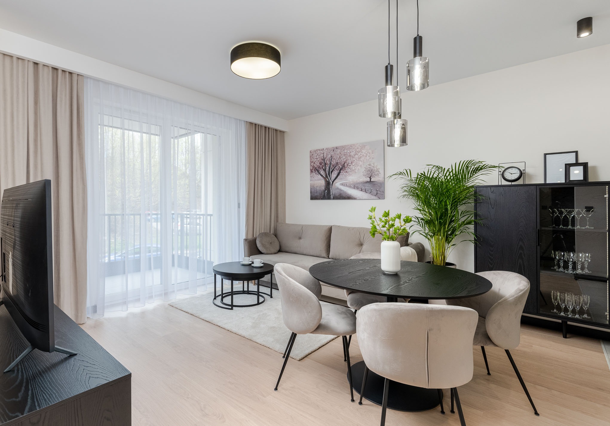 Jasne, nowocześnie urządzone wnętrze salonu z jadalnią: okrągły, czarny stół otoczony beżowymi krzesłami, sofa w odcieniach szarości, czarna szafka z szklanymi drzwiami i kieliszkami, obraz...