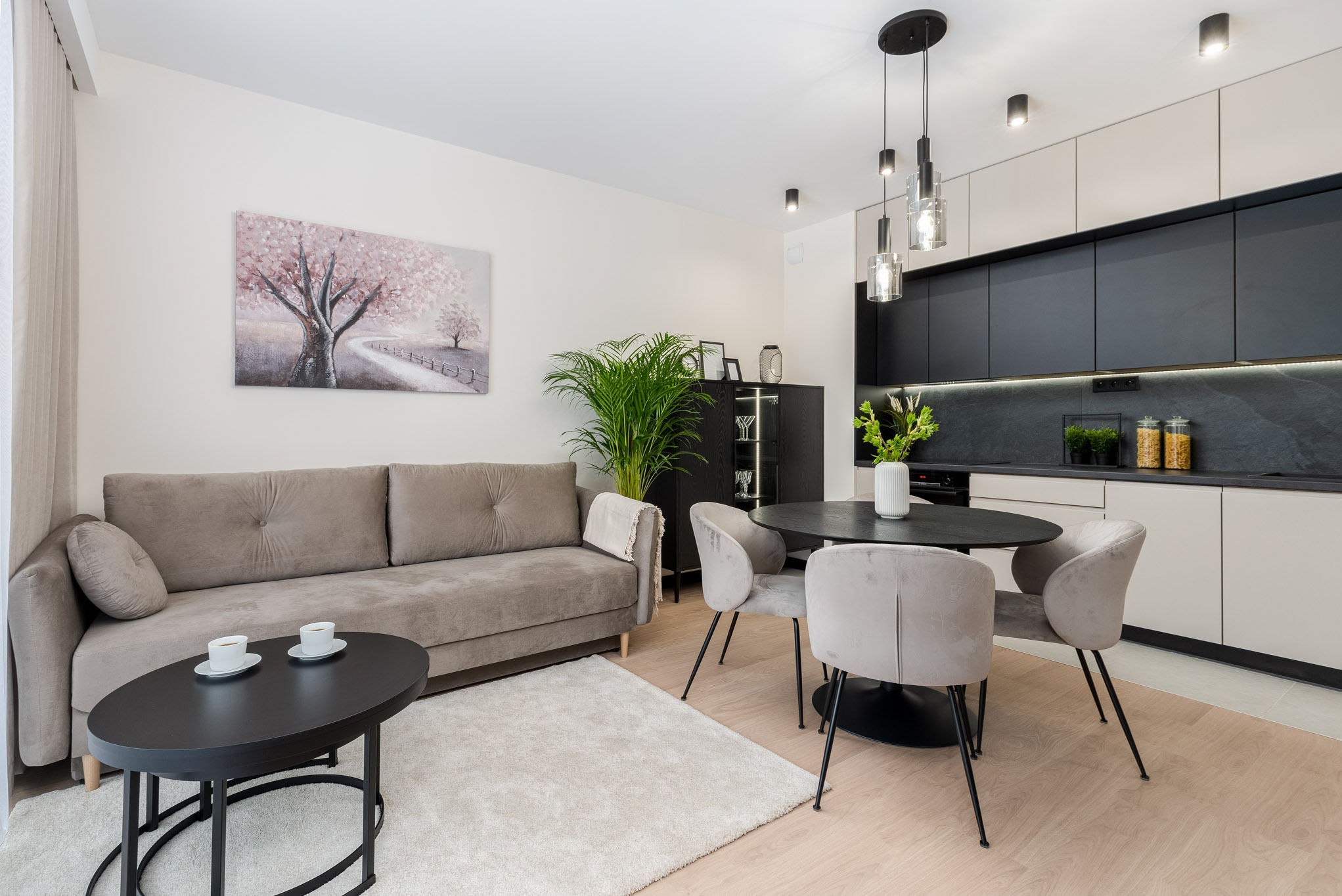 Jasne wnętrze salonu z aneksem kuchennym: szara sofa z poduszką, czarny stolik kawowy z dwoma filiżankami, okrągły stół z czterema krzesłami, zabudowa kuchenna z czarnymi frontami i jasnymi...