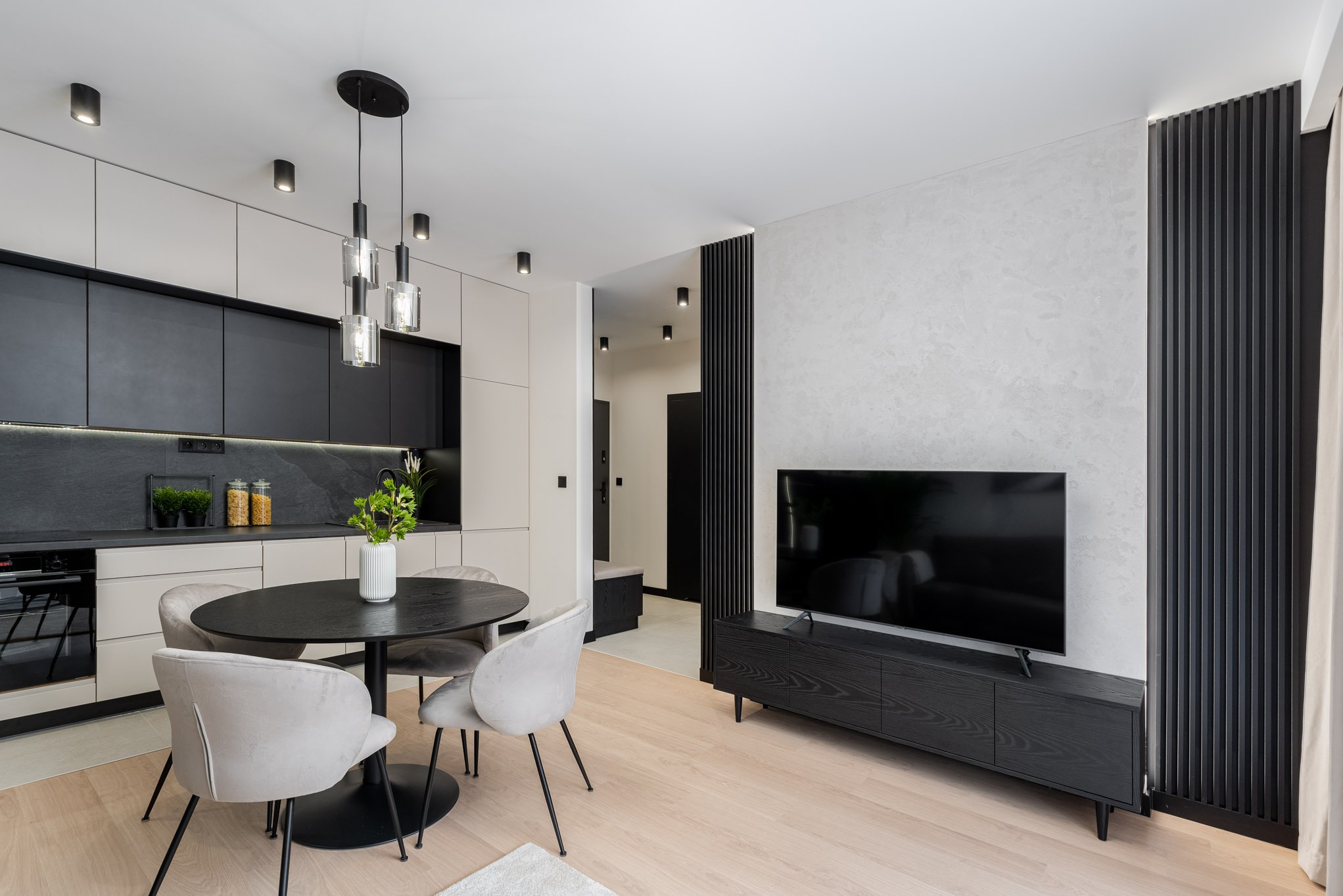 Nowoczesne wnętrze: czarna kuchnia z jasnymi szafkami, okrągły stół z welurowymi krzesłami, ściana z dekoracyjnymi lamelami, telewizor na szafce RTV.