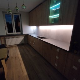 Vibe Wood - Nowoczesna kuchnia z drewnianymi frontami, podświetleniem LED pod szafkami wiszącymi i przeszkloną szafką z turkusowym oświetleniem, widok na fragment blatu z wbudowaną płytą grzewczą.