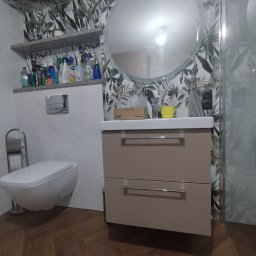Vibe Wood - Nowoczesna łazienka z szafką pod umywalką w kolorze beżowym, okrągłym lustrem i toaletą podwieszaną na tle ściany z motywem roślinnym.