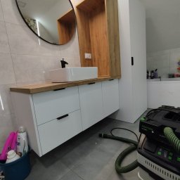 Vibe Wood - Nowoczesna łazienka z umywalką nablatową na szafce z białymi frontami i drewnianym blatem, okrągłe lustro w czarnej ramie, widoczny odkurzacz przemysłowy Festool na podłodze.