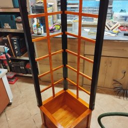 Vibe Wood - Kątowa drewniana konstrukcja z donicą u podstawy, czarne pionowe elementy i pomarańczowe poprzeczki, widoczna w warsztacie z narzędziami w tle.