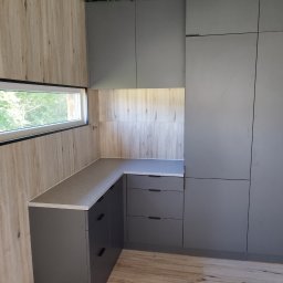 Vibe Wood - Szara zabudowa kuchenna z blatem, widoczna z perspektywy narożnika pomieszczenia, z dekoracyjną zielenią na suficie i oknem z widokiem na zieleń na ścianie.