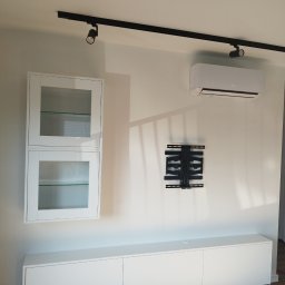 Vibe Wood - Biała zabudowa meblowa pod telewizor z szafkami wiszącymi z przeszklonymi drzwiczkami, zamontowany klimatyzator i uchwyt do telewizora na ścianie, oświetlenie szynowe.