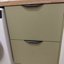 Vibe Wood - Szafka pod umywalkę z dwoma szufladami w odcieniach oliwkowej zieleni, z blatem imitującym drewno, czarne, minimalistyczne uchwyty, obok pralka.