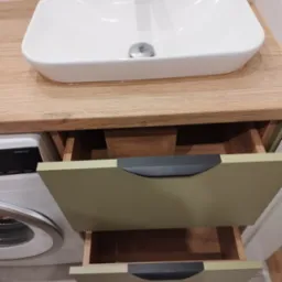 Biała, prostokątna umywalka nablatowa na blacie z jasnego drewna, poniżej szafka z dwoma wysuwanymi szufladami w kolorze oliwkowym, obok pralka.