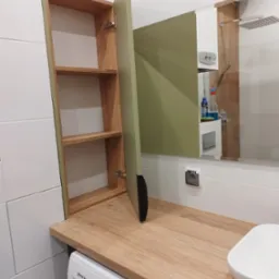 Łazienka z szafką z otwartymi półkami i zielonymi drzwiczkami, blat z drewnopodobnego laminatu nad pralką, umywalka w rogu.