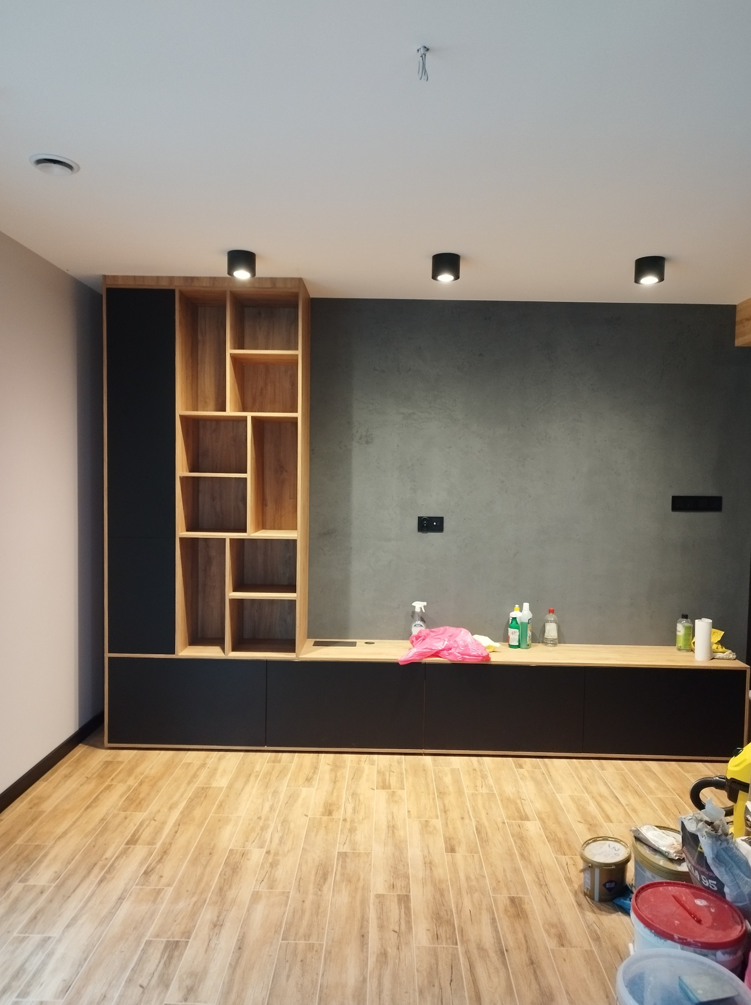 Częściowo ukończona zabudowa meblowa z czarnymi frontami i regałem z jasnego drewna, widoczne narzędzia i środki czystości sugerują prace wykończeniowe.