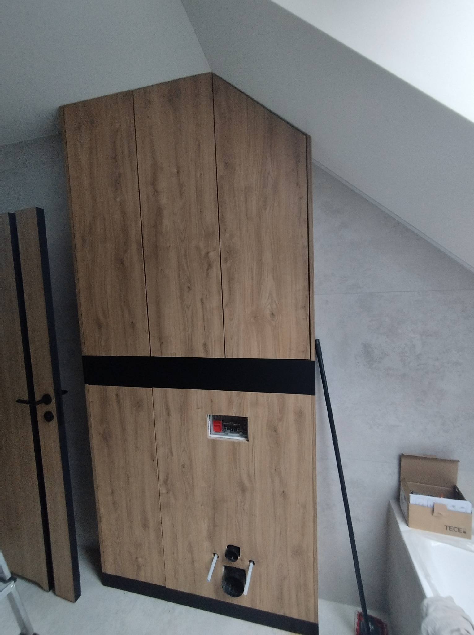 Wnęka zabudowana drewnopodobną szafą z czarnym pasem, widoczne przyłącza hydrauliczne, w tle skośny sufit i płytki ścienne, obok drzwi z podobnym wykończeniem.