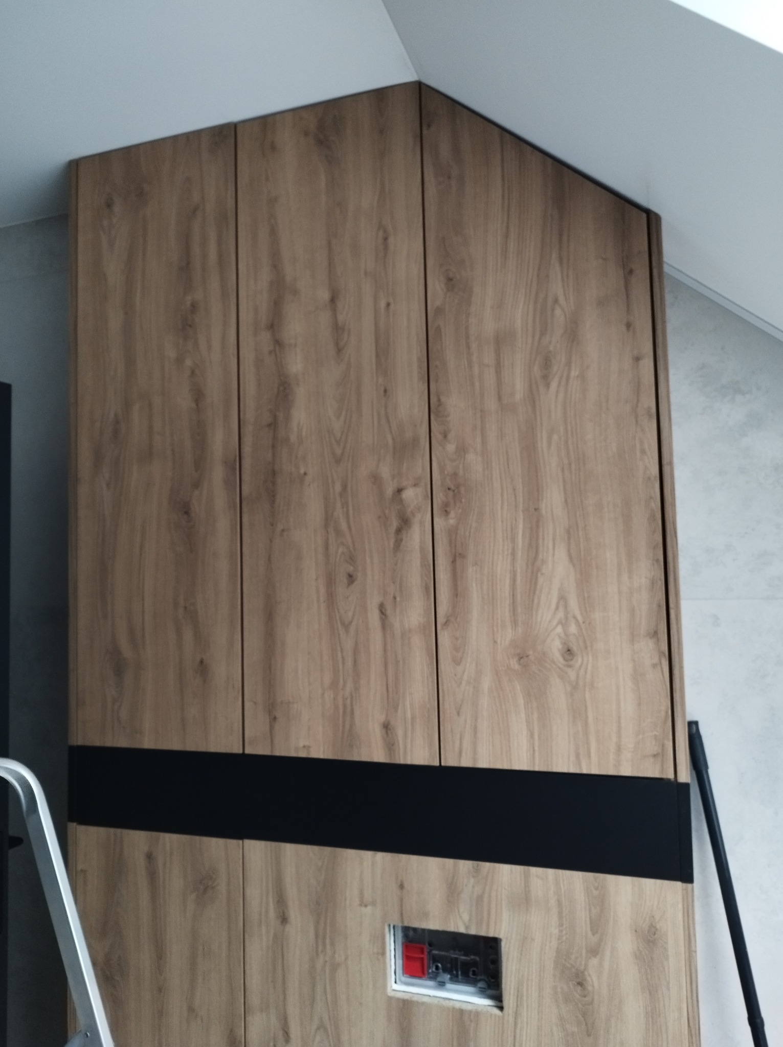 Szafa wnękowa z drewnopodobnymi frontami, czarnym pasem dekoracyjnym i otworem instalacyjnym w dolnej części, dopasowana do skosu dachu.