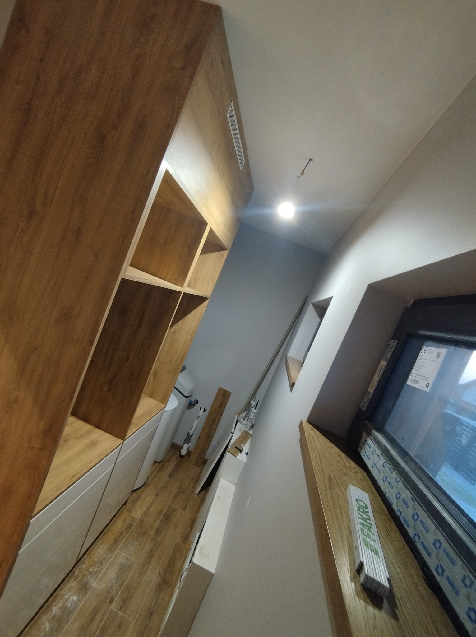 Drewniana zabudowa wnęki z półkami i białymi szafkami, widok z dołu, fragment okna z miarką Fakro, białe ściany i drewniana podłoga.