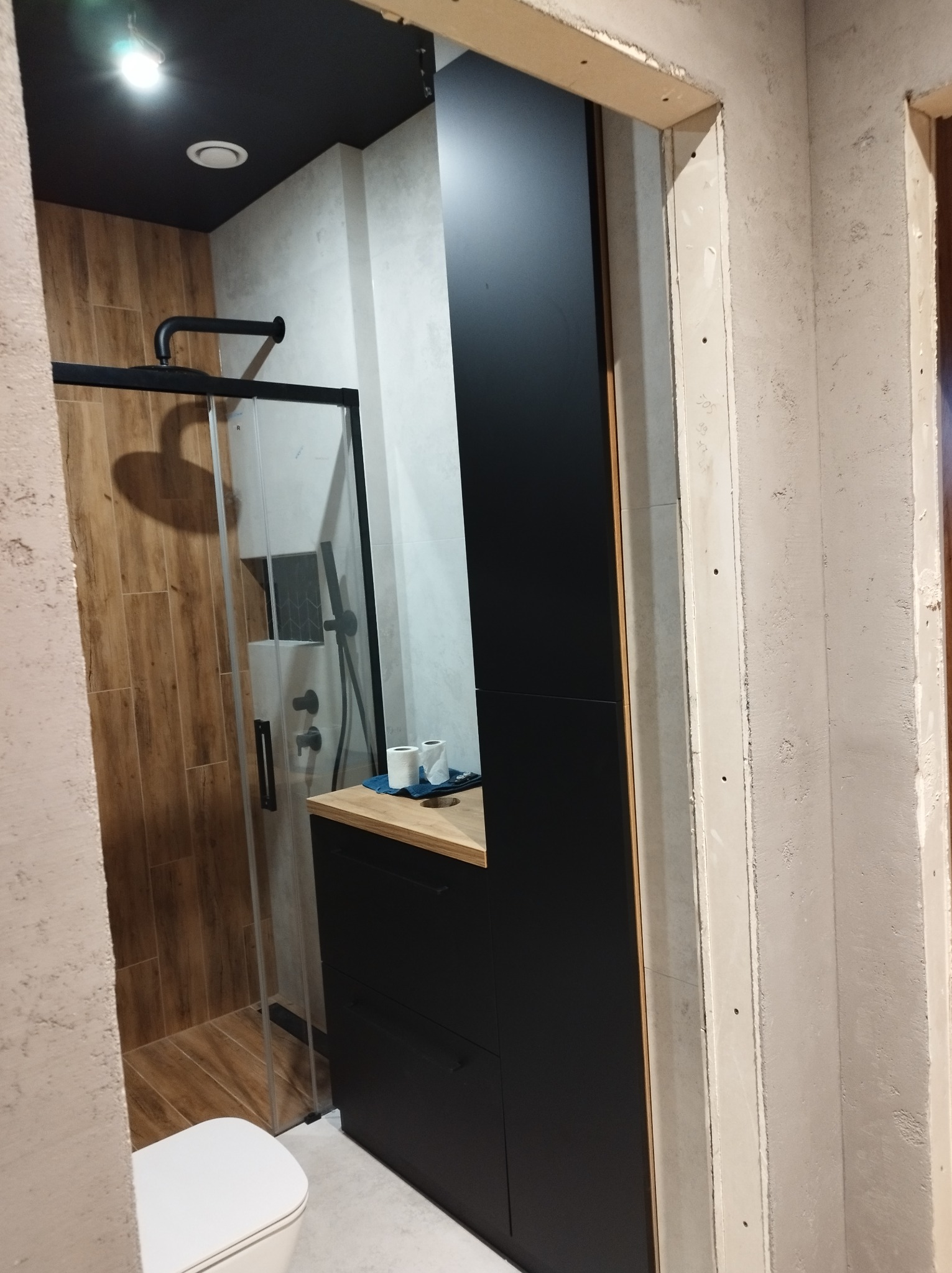 Nowoczesna łazienka z kabiną prysznicową typu walk-in, czarną szafką z drewnianym blatem, wykończona w drewnie i szarości, widok z surowego otworu drzwiowego.