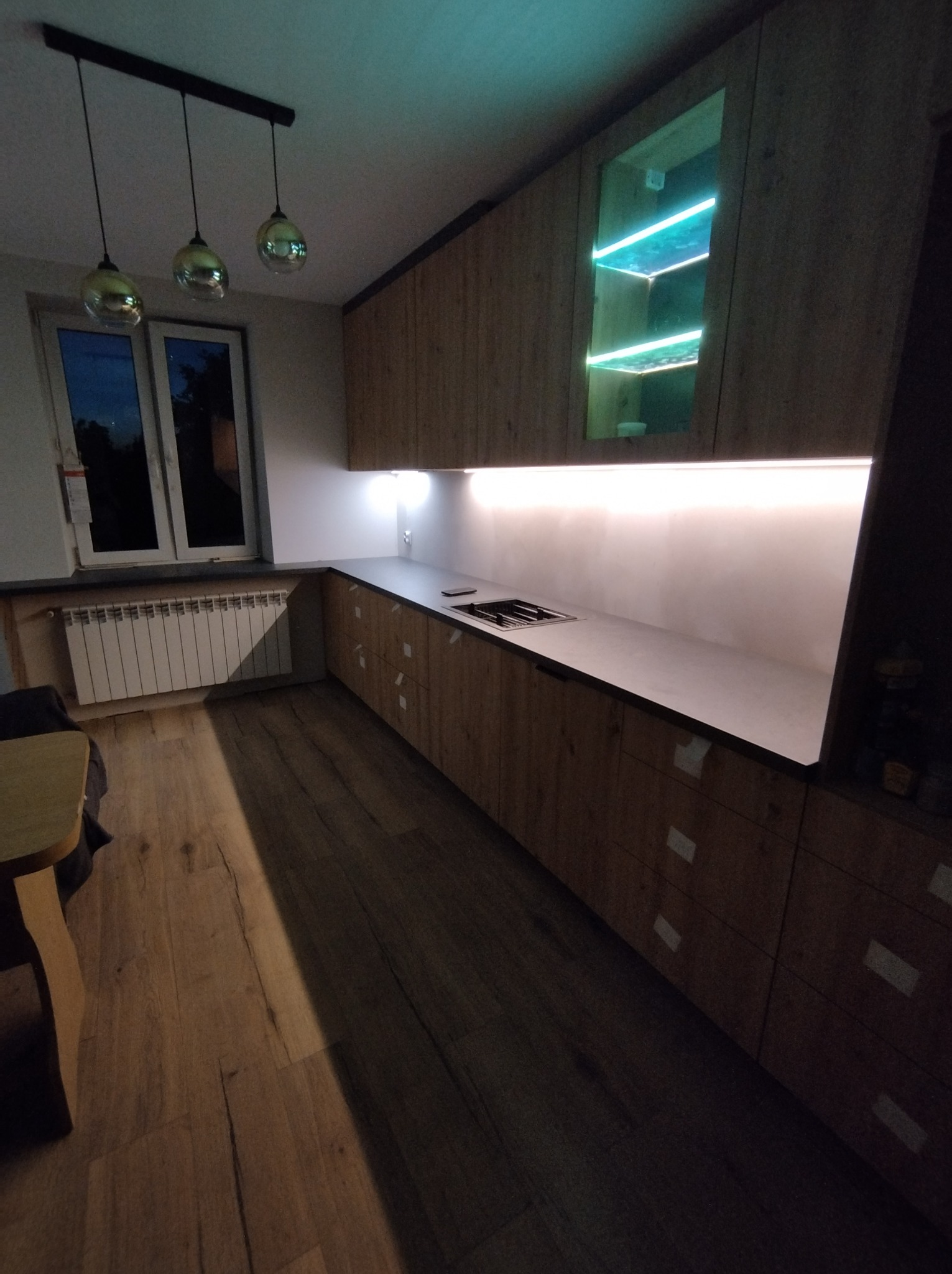 Nowoczesna kuchnia z drewnianymi frontami, podświetleniem LED pod szafkami wiszącymi i przeszkloną szafką z turkusowym oświetleniem, widok na fragment blatu z wbudowaną płytą grzewczą.