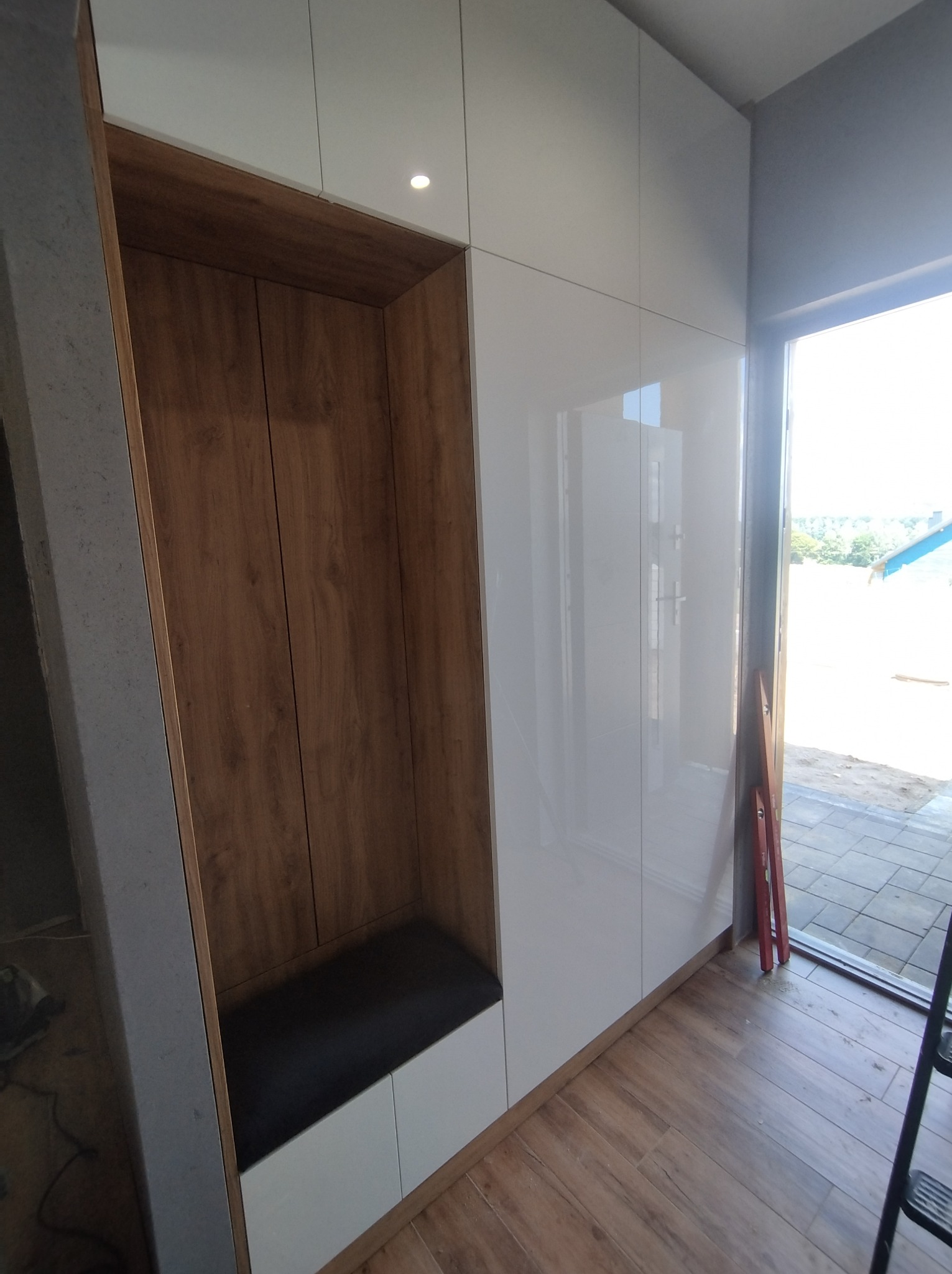 Nowoczesna szafa wnękowa z drewnianą wnęką i siedziskiem, białe lakierowane fronty, widoczny fragment okna i podłogi.