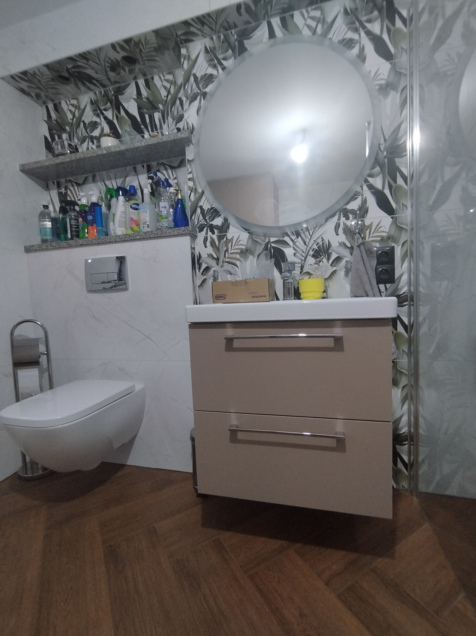 Nowoczesna łazienka z szafką pod umywalką w kolorze beżowym, okrągłym lustrem i toaletą podwieszaną na tle ściany z motywem roślinnym.