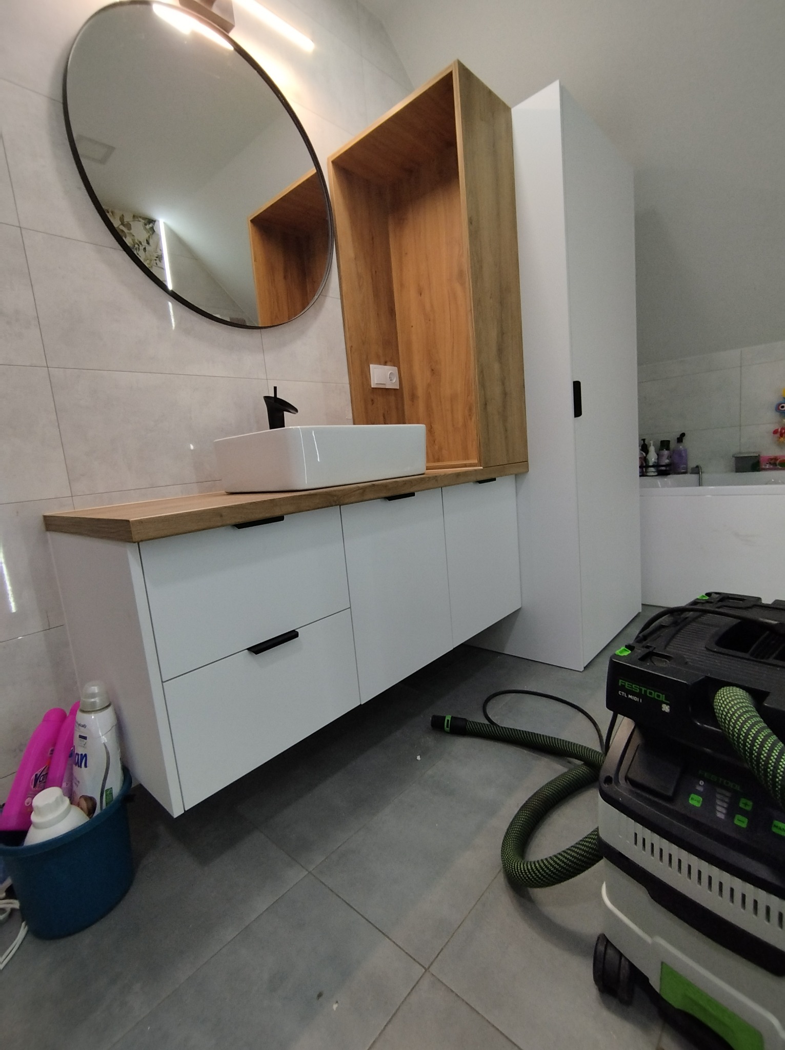 Nowoczesna łazienka z umywalką nablatową na szafce z białymi frontami i drewnianym blatem, okrągłe lustro w czarnej ramie, widoczny odkurzacz przemysłowy Festool na podłodze.