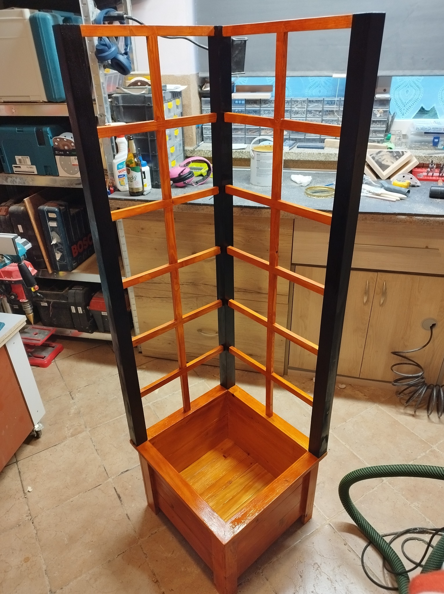 Kątowa drewniana konstrukcja z donicą u podstawy, czarne pionowe elementy i pomarańczowe poprzeczki, widoczna w warsztacie z narzędziami w tle.