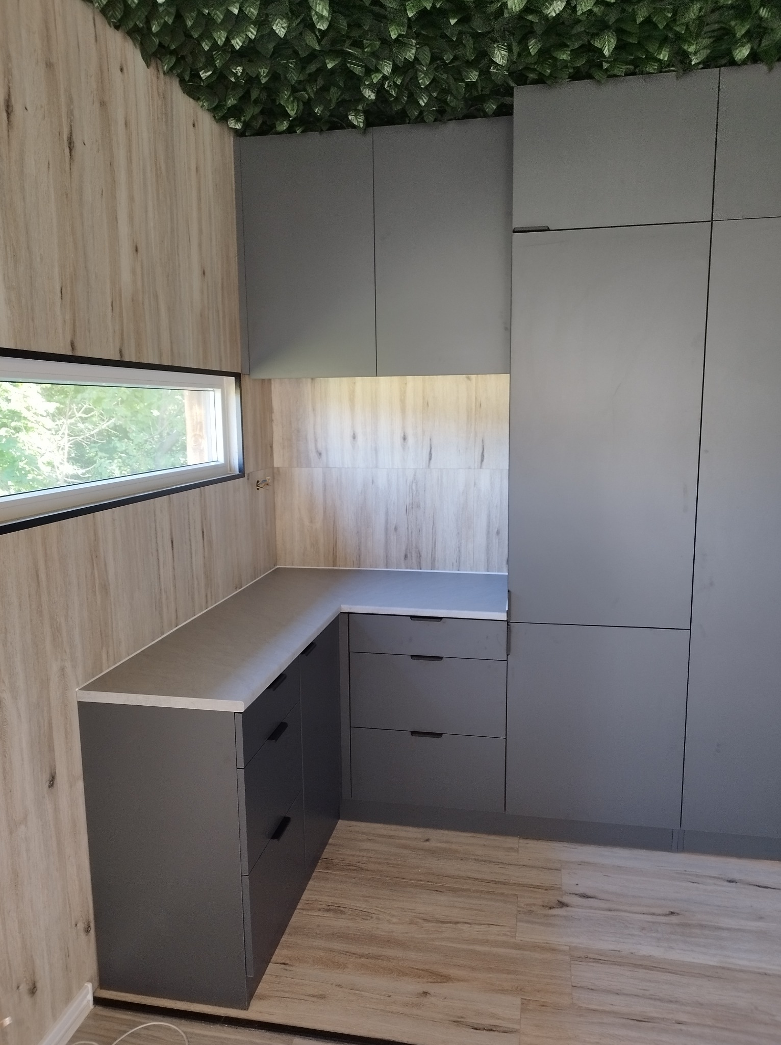 Szara zabudowa kuchenna z blatem, widoczna z perspektywy narożnika pomieszczenia, z dekoracyjną zielenią na suficie i oknem z widokiem na zieleń na ścianie.