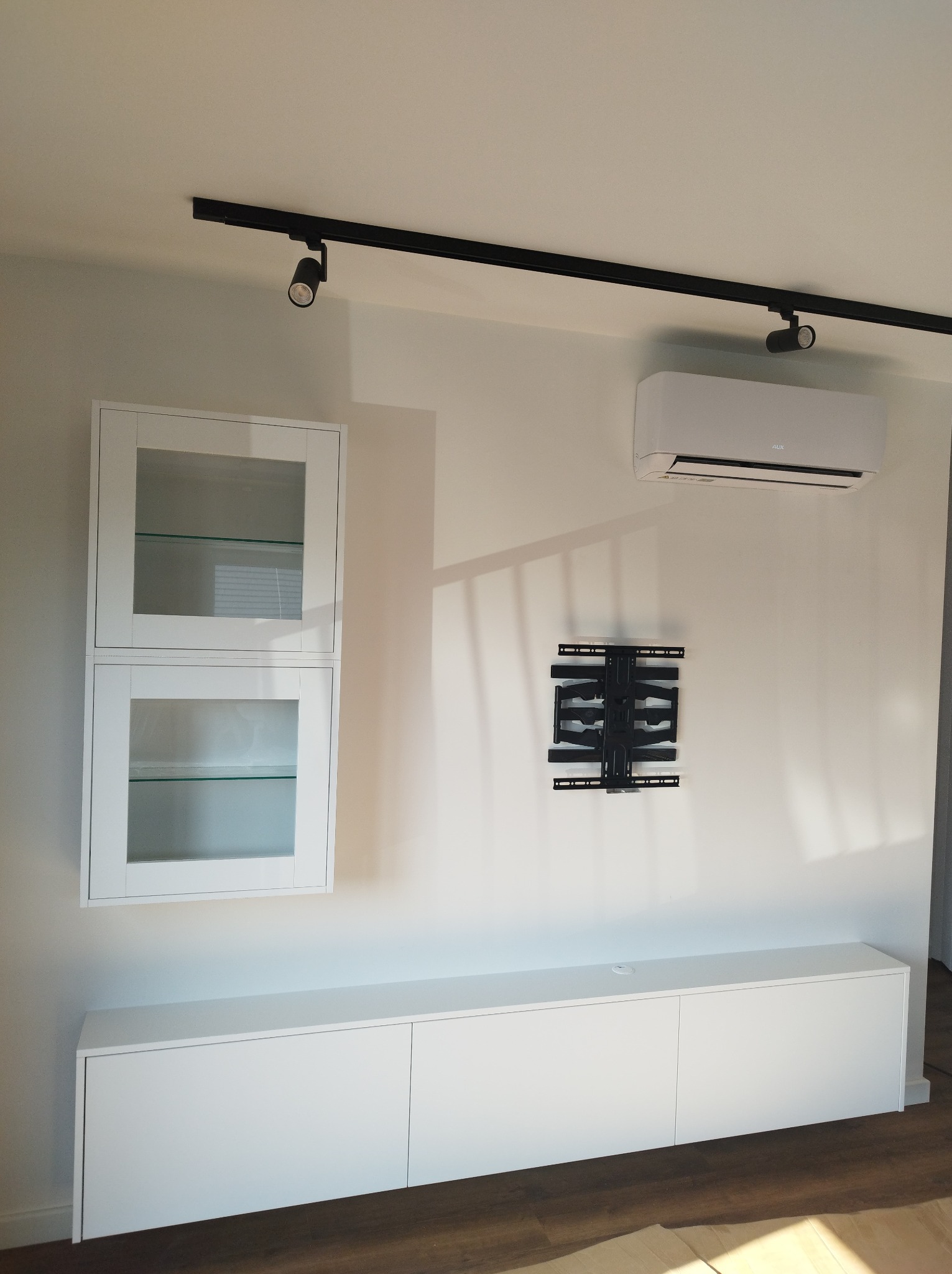 Biała zabudowa meblowa pod telewizor z szafkami wiszącymi z przeszklonymi drzwiczkami, zamontowany klimatyzator i uchwyt do telewizora na ścianie, oświetlenie szynowe.