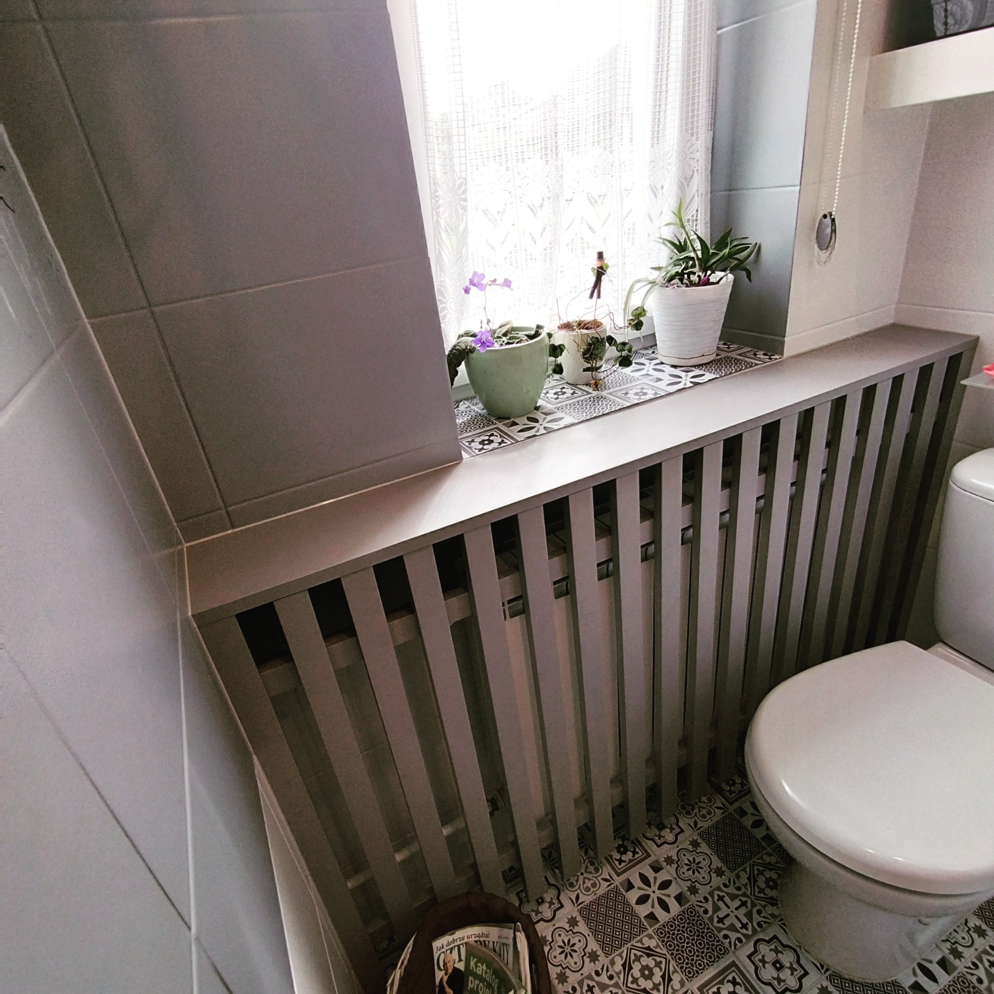 Szara zabudowa grzejnika w łazience z kwiatami w doniczkach na parapecie i wzorzystą podłogą.
