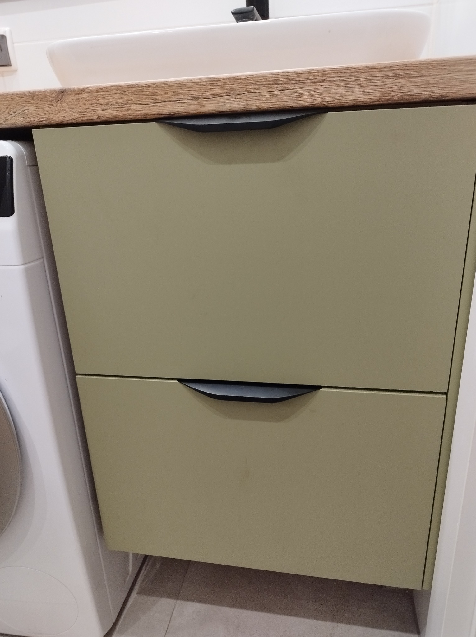 Szafka pod umywalkę z dwoma szufladami w odcieniach oliwkowej zieleni, z blatem imitującym drewno, czarne, minimalistyczne uchwyty, obok pralka.