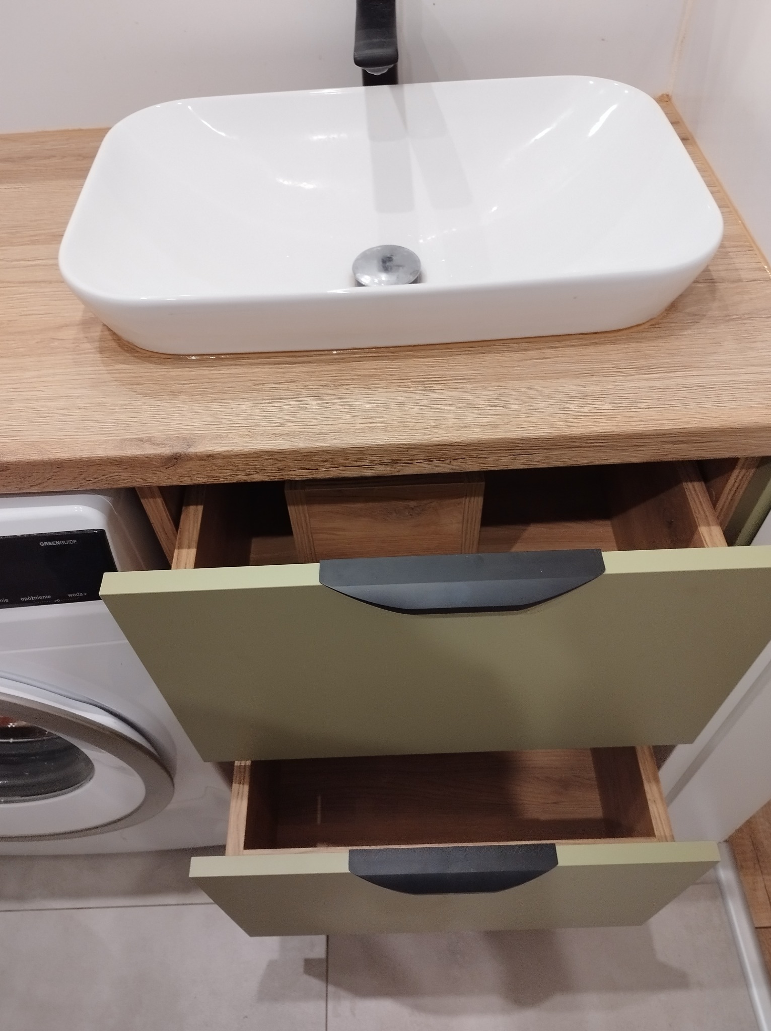 Biała, prostokątna umywalka nablatowa na blacie z jasnego drewna, poniżej szafka z dwoma wysuwanymi szufladami w kolorze oliwkowym, obok pralka.