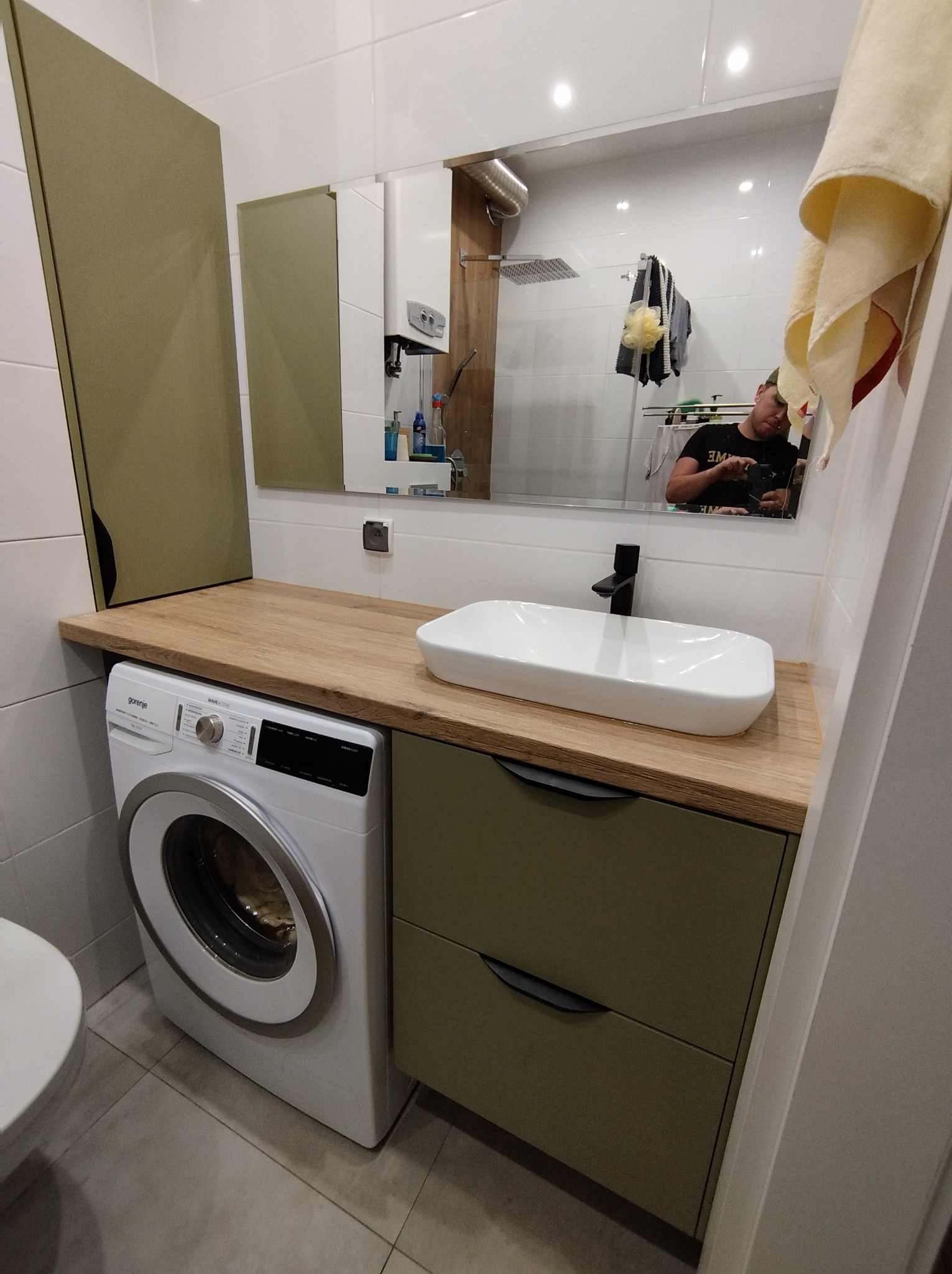 Nowoczesna łazienka z zabudową na wymiar w odcieniach bieli i oliwki, z pralką wkomponowaną pod blatem umywalki i dużym lustrem odbijającym fragment kabiny prysznicowej.