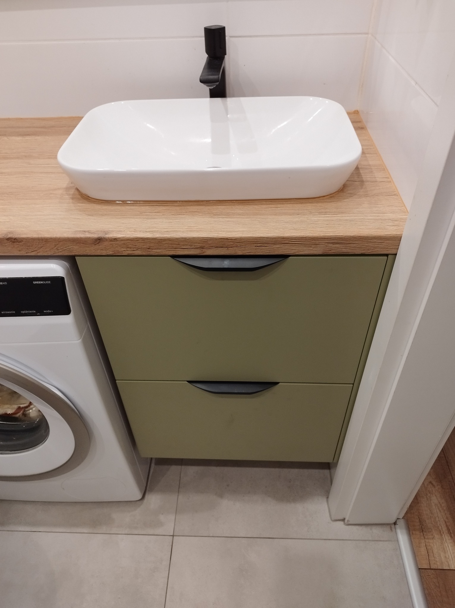 Nowoczesna łazienka z umywalką nablatową, drewnianym blatem i szafką w kolorze oliwkowym, obok pralka.